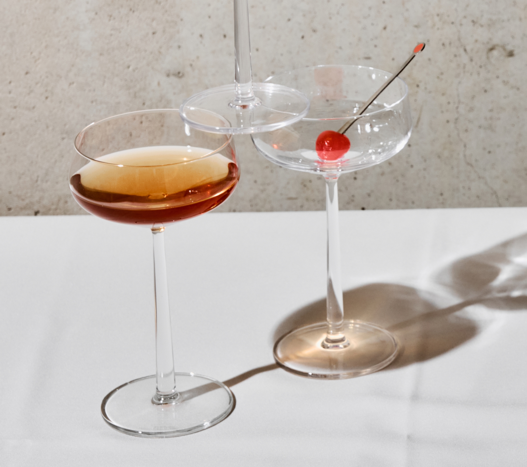 Viisi kuppilasia, joissa on meripihkanvärisiä cocktaileja ja kirsikoita, heittävät pitkiä varjoja valkoiselle pöydälle betoniseinää vasten.