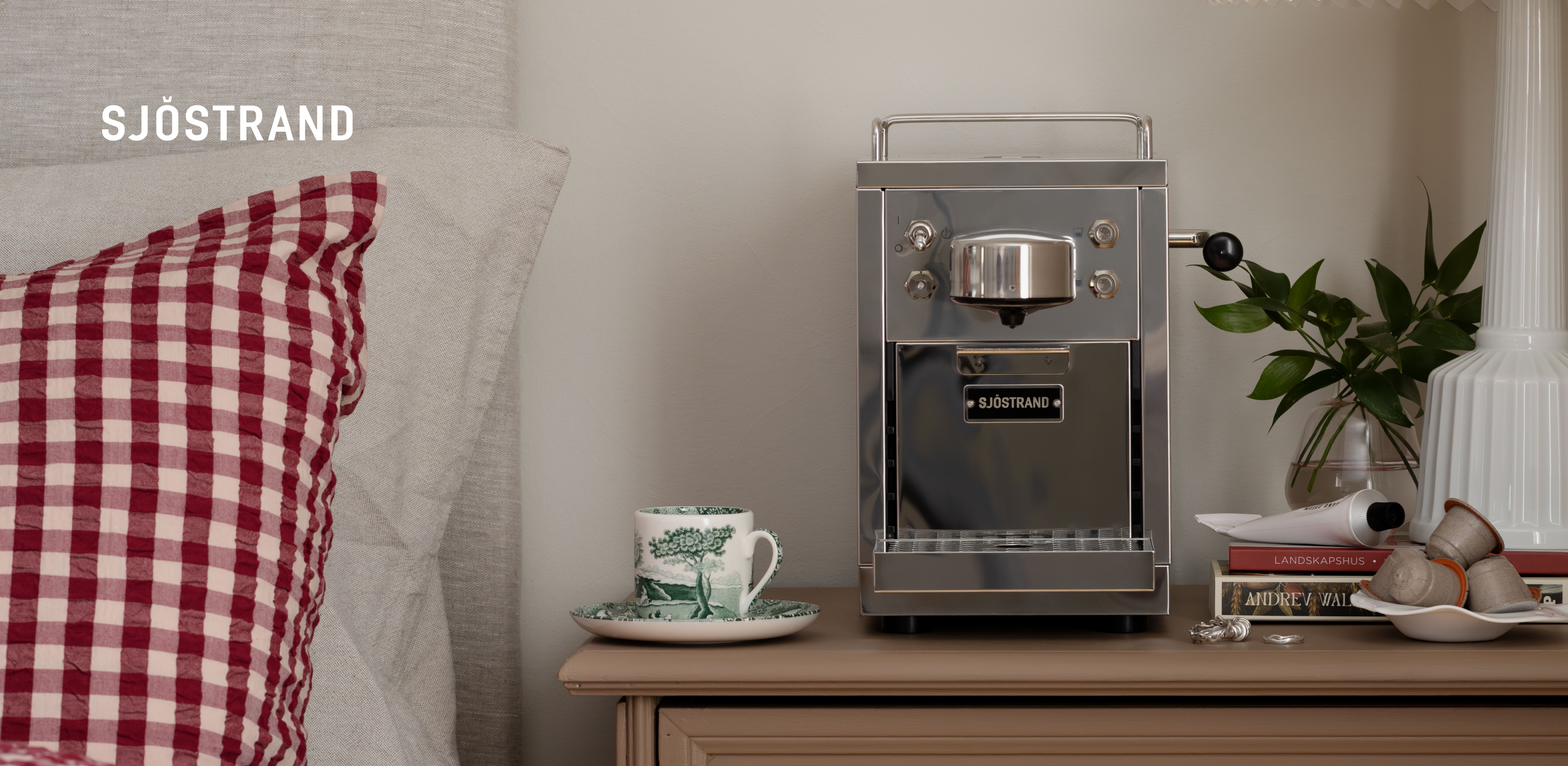 En silverfärgad Sjöstrand-kaffemaskin, en liten mönstrad kopp, böcker och kaffekapslar på ett nattduksbord, bredvid en röd kudde..