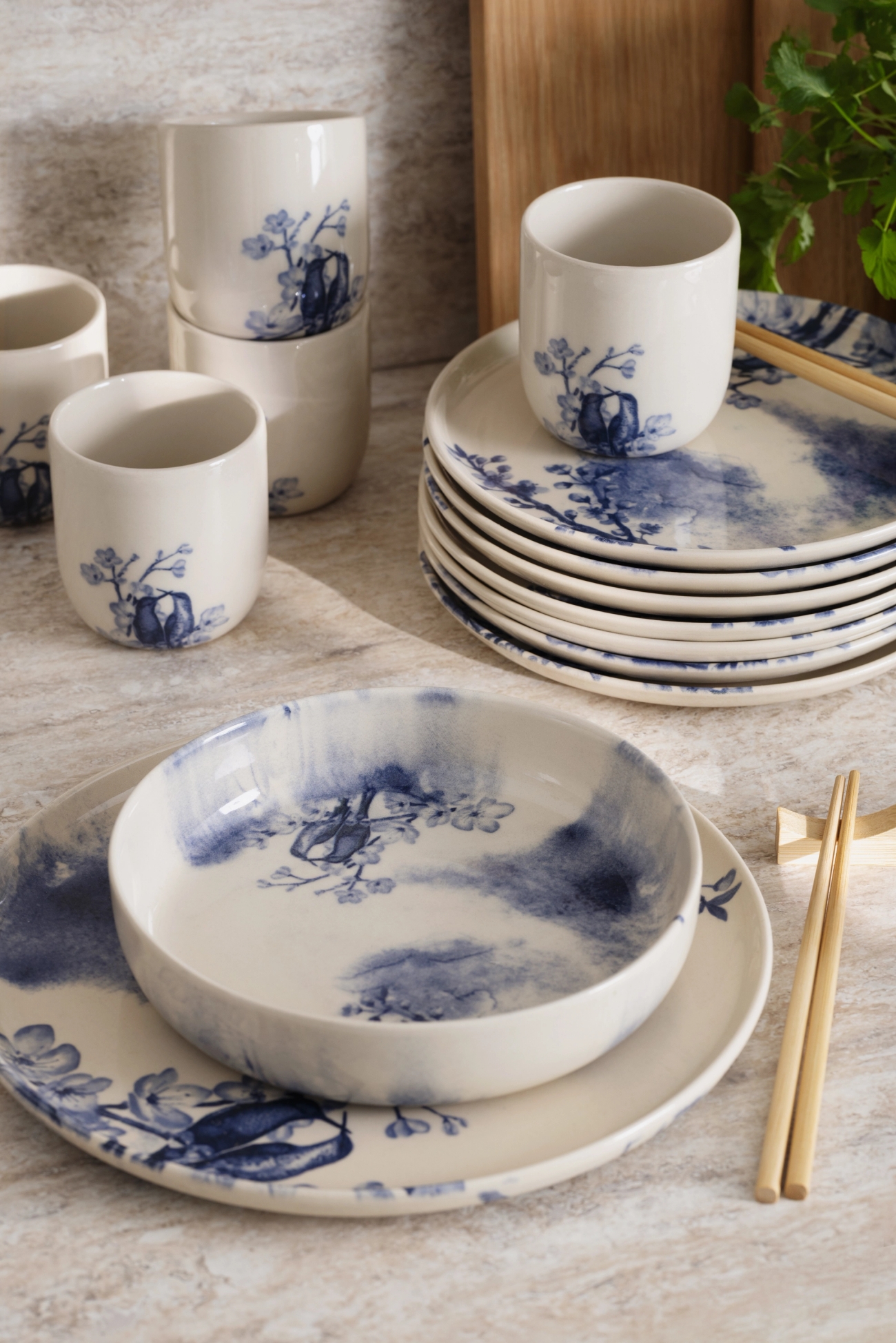 Hvitt keramisk servise med blå blomster- og akvarelldesign, inkludert kopper, stablede tallerkener og en bolle med spisepinner.