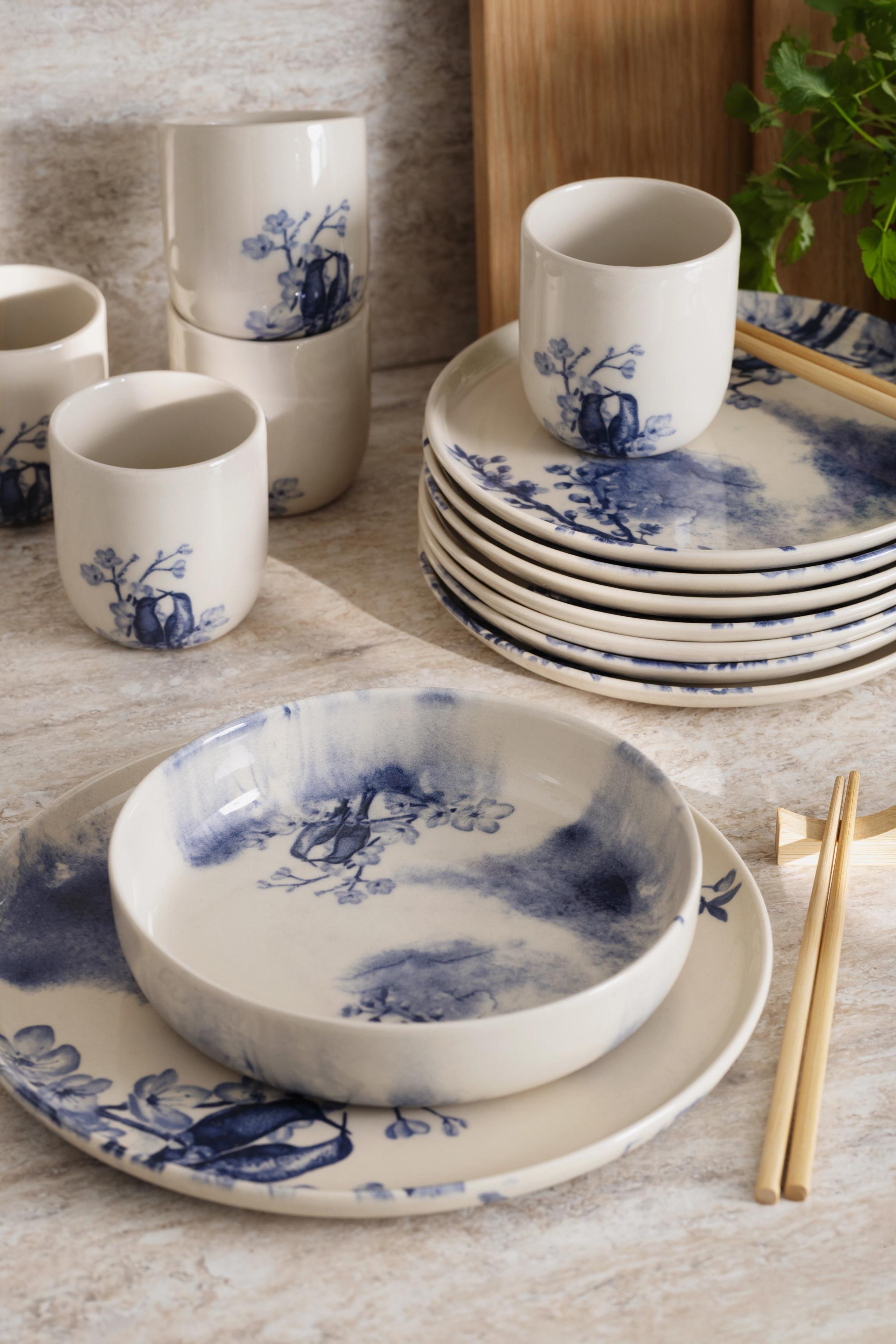 Vitt keramiskt porslin med blå akvarellfärgade fågel- och blommönster, inklusive koppar, staplade tallrikar, en skål och ätpinnar.