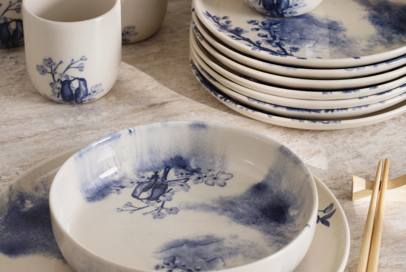 En kollektion av vitt keramiskt porslin med blå botaniska mönster och abstrakta blå stänk, inklusive tallrikar, skålar och koppar med ätpinnar.