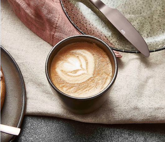 Kaffemugg utan öra med latteart, placerad på dukad frukostdukning med bestick och tallrik i naturliga färger.