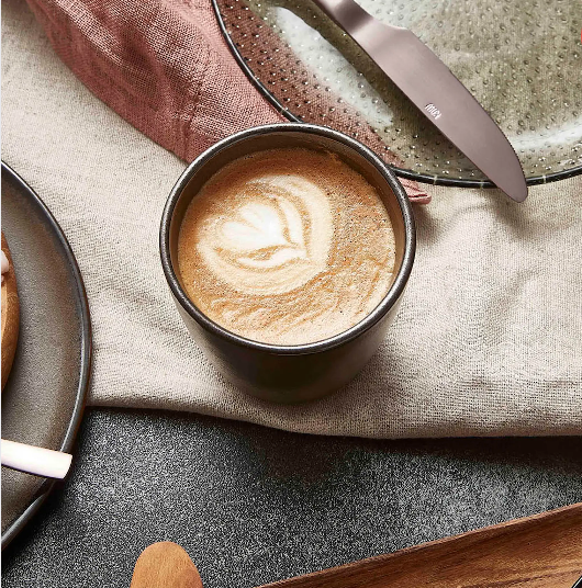 Kaffemugg utan öra med latteart, placerad på dukad frukostdukning med bestick och tallrik i naturliga färger.