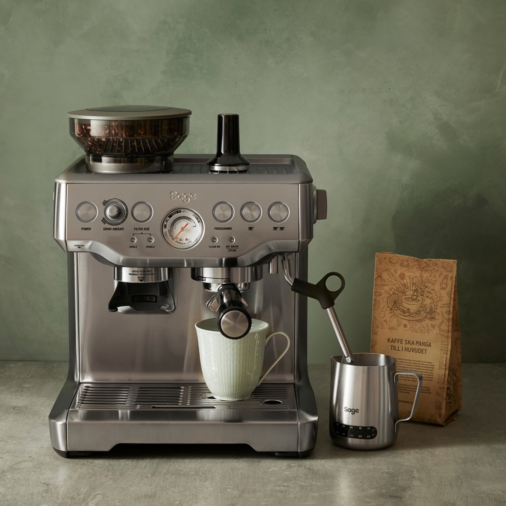 Ruostumattomasta teräksestä valmistettu espressokone jauhaa kahvipapuja ja valmistaa kahvia vihreään mukiin, vieressä maitokannu ja kahvipapupussi.