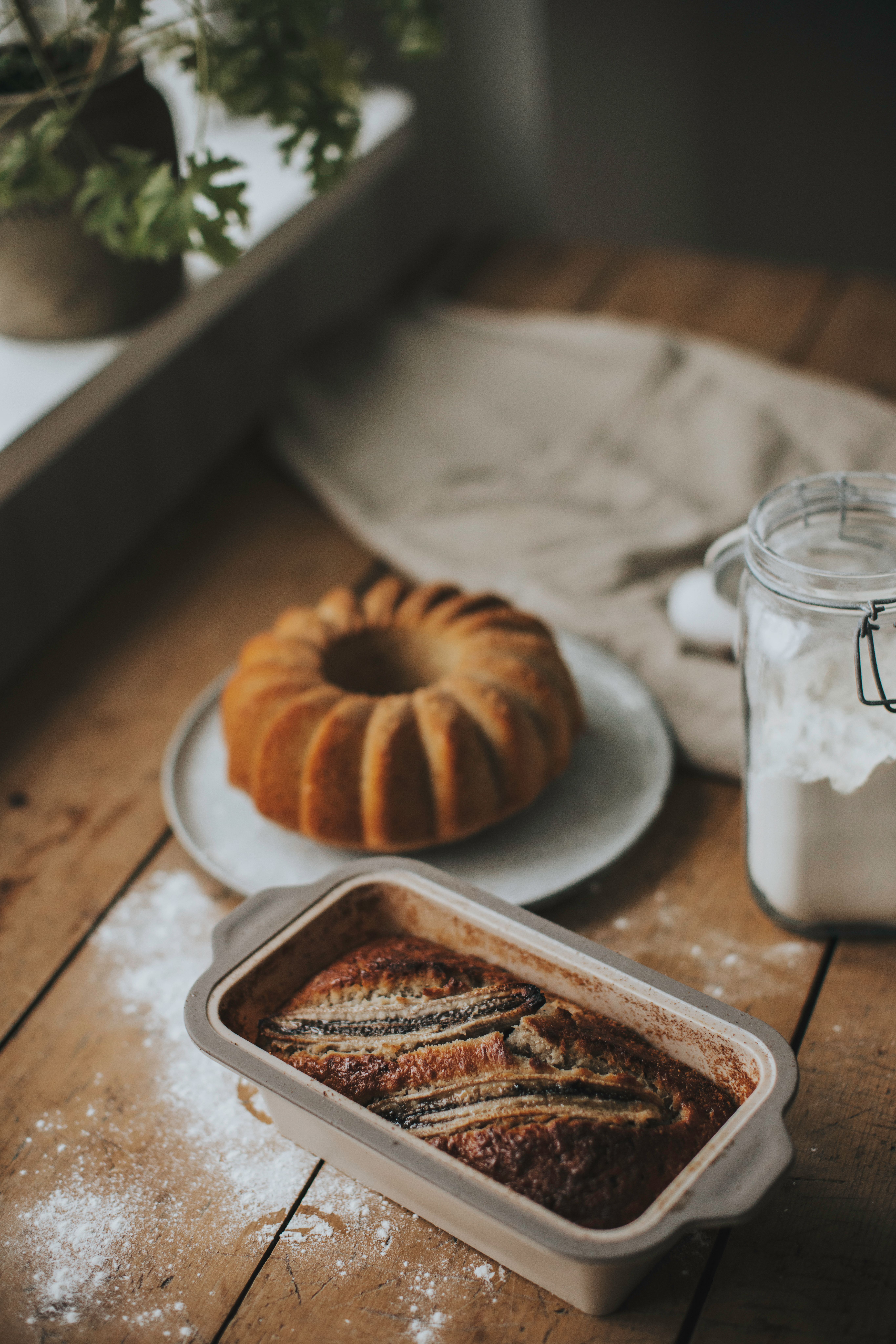 Nybakat bananbröd i en brödform, med en Bundtkaka och en burk mjöl i bakgrunden.