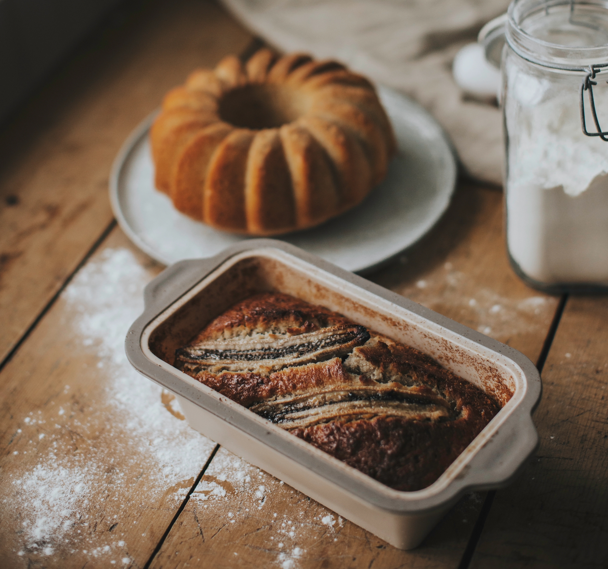 Nybakat bananbröd i en brödform, med en Bundtkaka och en burk mjöl i bakgrunden.
