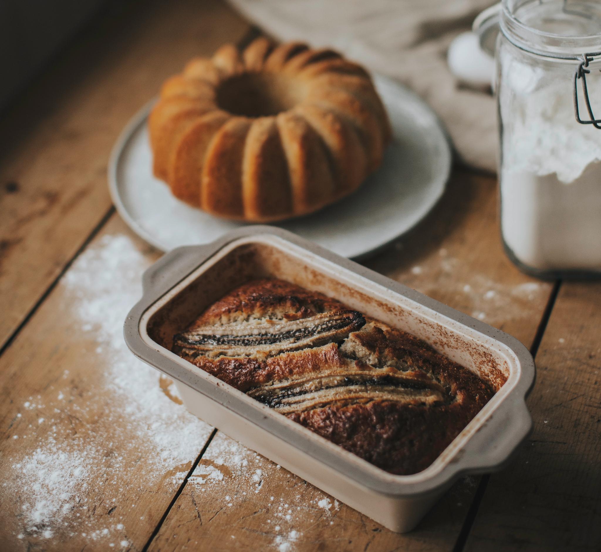 Nybakat bananbröd i en brödform, med en Bundtkaka och en burk mjöl i bakgrunden.