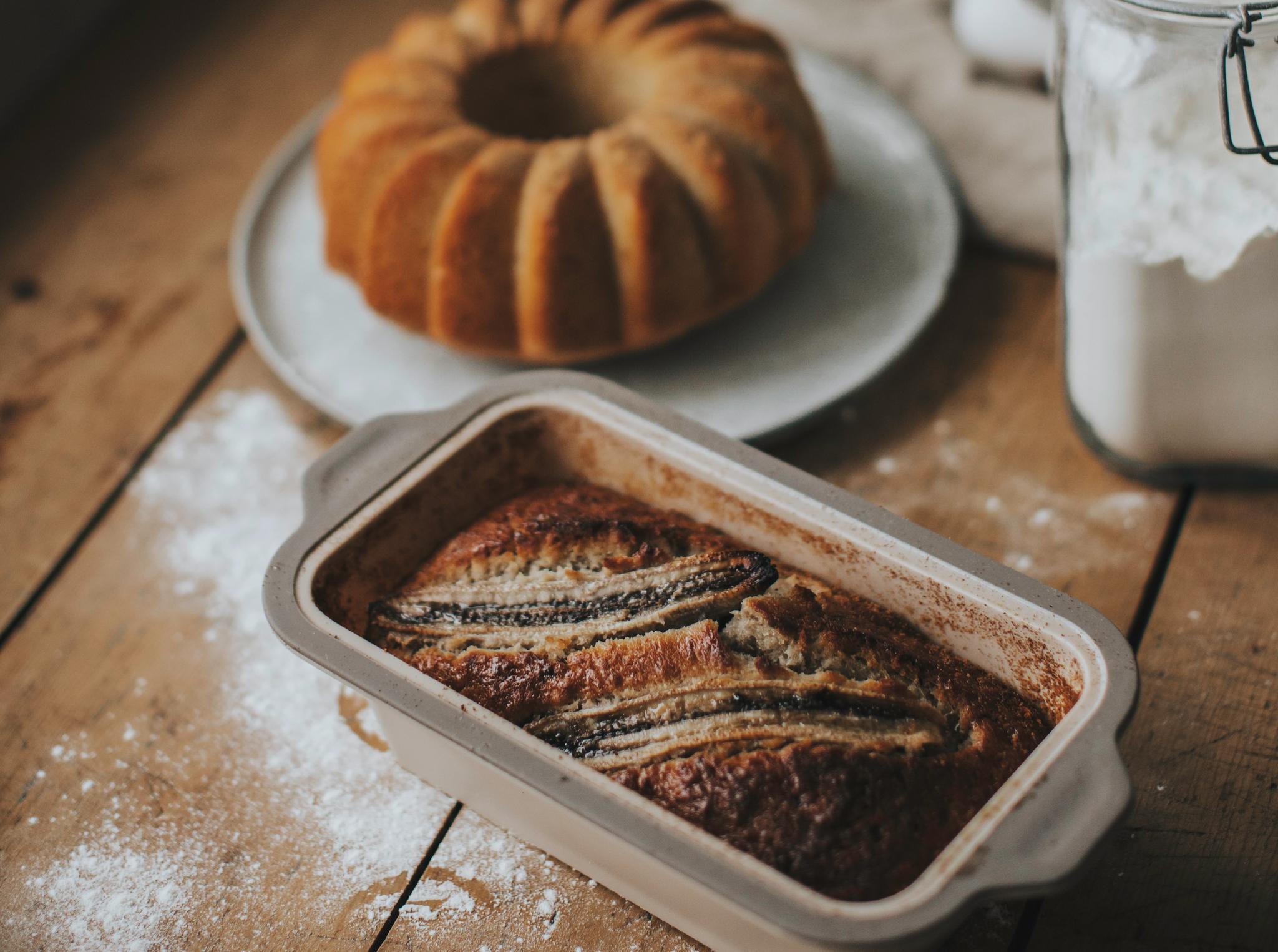 Nystekt bananbrød og formkake står på et rustikt trebord med mel drysset rundt.