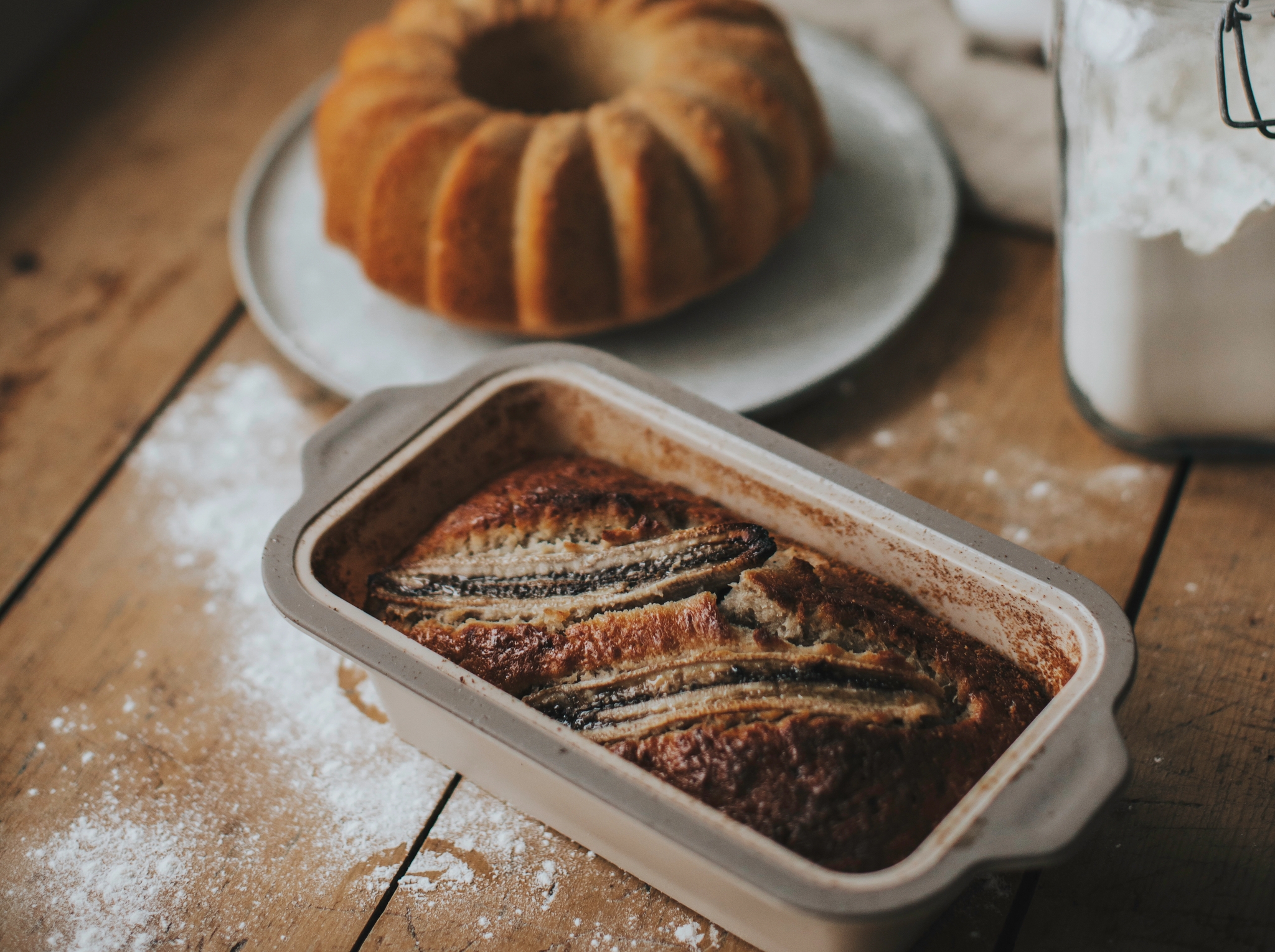 Nystekt bananbrød og formkake står på et rustikt trebord med mel og bakeutstyr rundt.