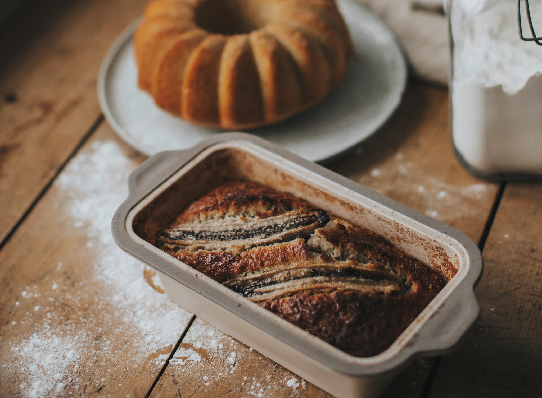 Nybakat bananbröd i form på ett rustikt köksbord med sockerkaka och mjöl i bakgrunden.