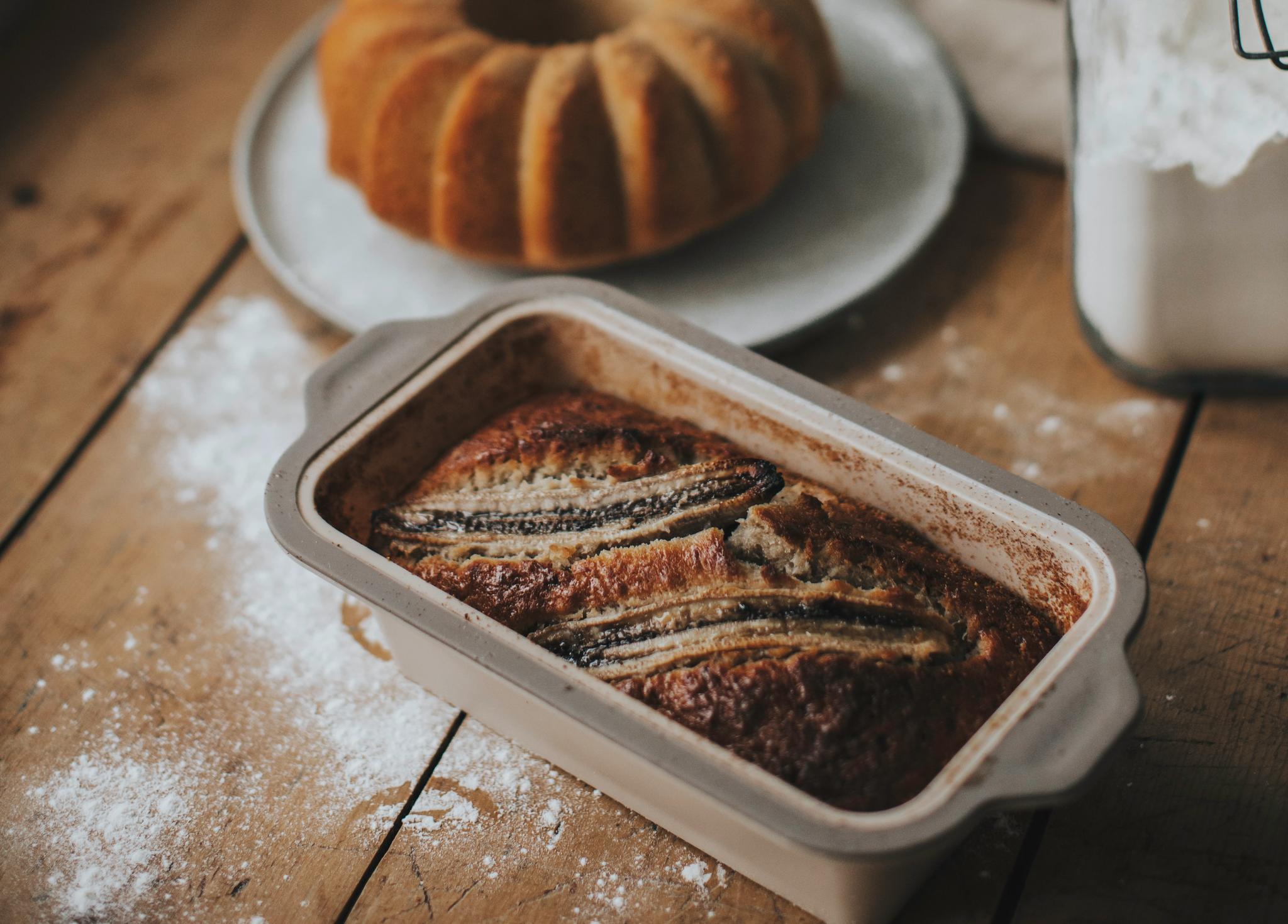 Nygräddad banankaka i form på ett rustikt träbord med sockerkaka och mjölburk i bakgrunden.