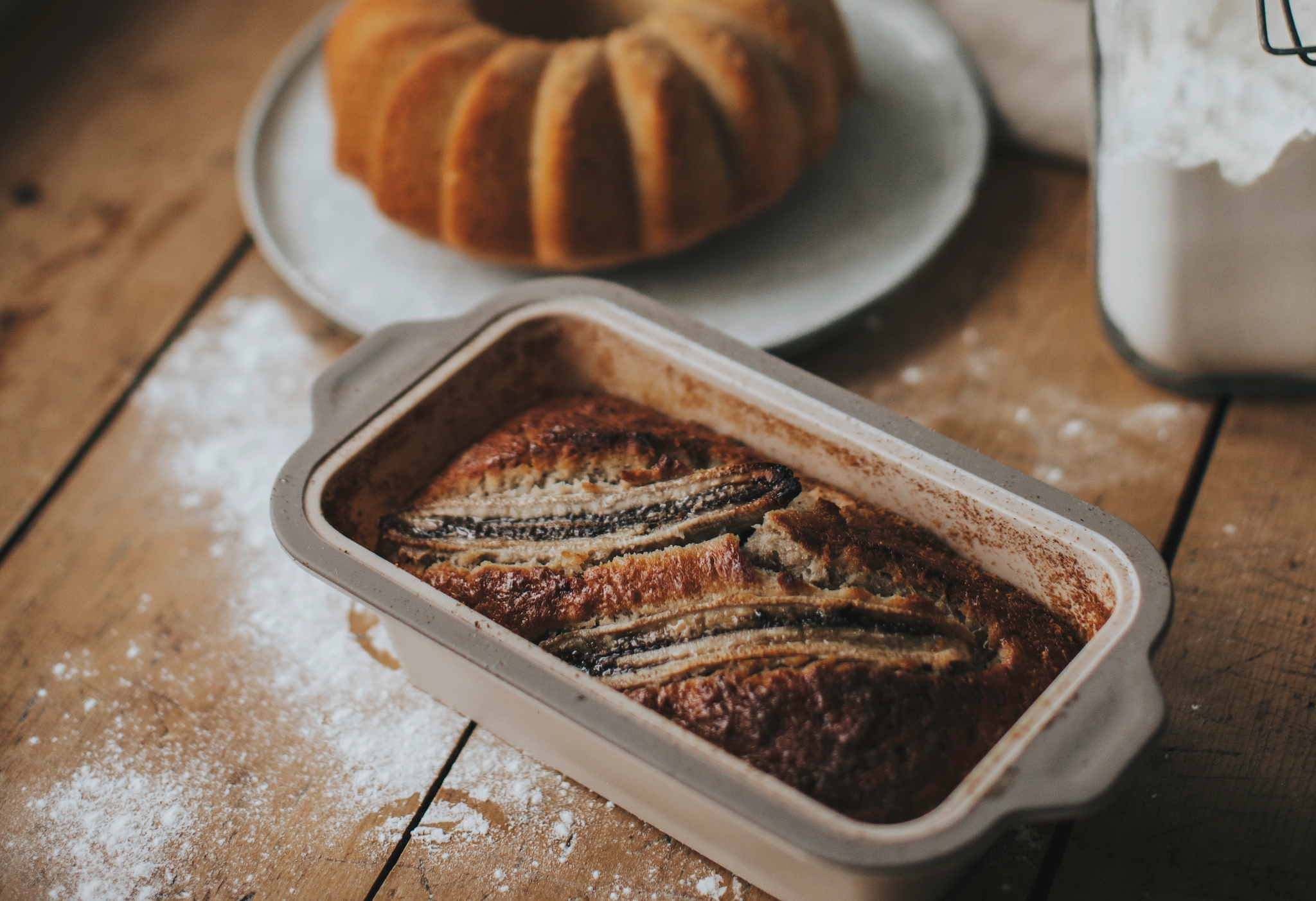 Nygräddad banankaka i form på ett rustikt träbord med mjöl, sockerkaka och gröna växter i bakgrunden.