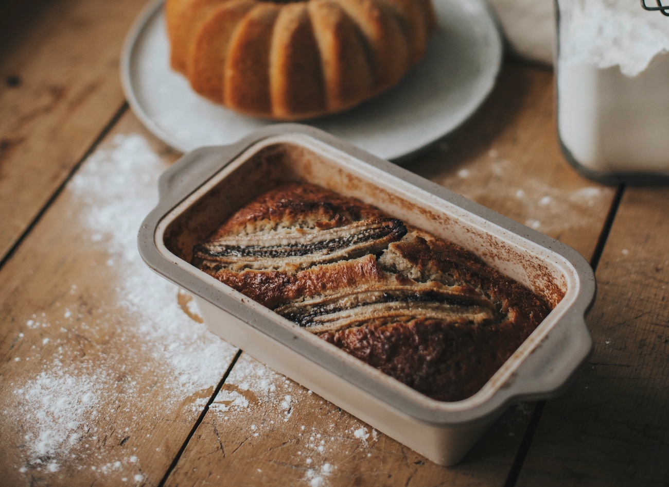 Et bananbrød med bananskiver i en panne, en bundtkake på en tallerken og bakeingredienser på et rustikt trebord.