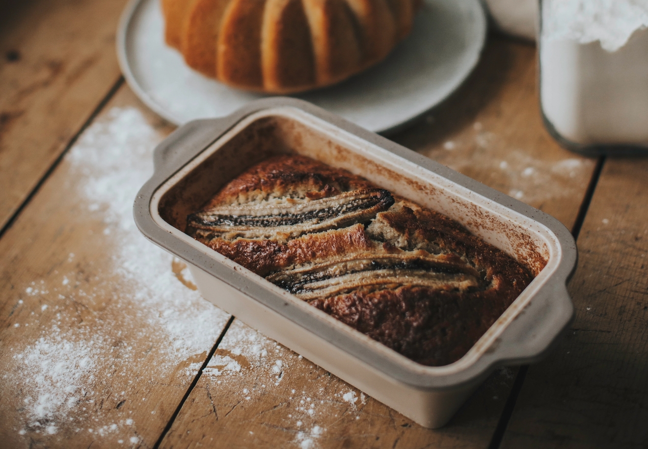 Et bananbrød med bananskiver i en panne, en bundtkake på en tallerken og bakeingredienser på et rustikt trebord.
