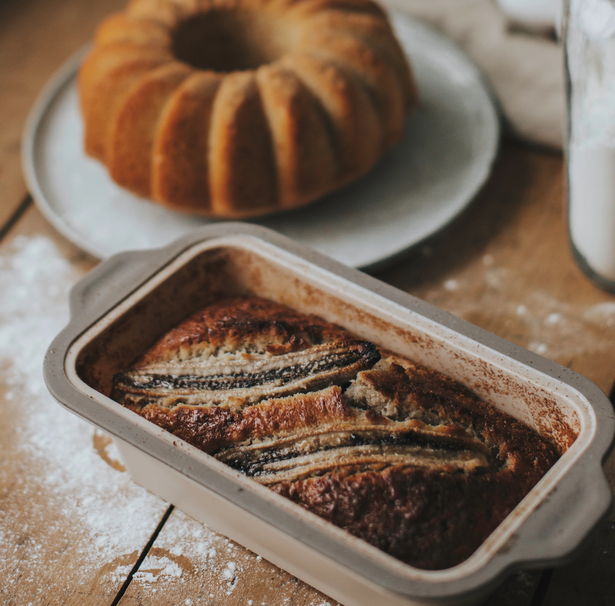 Nygräddad banankaka i form på ett rustikt träbord med sockerkaka och mjölburk i bakgrunden.