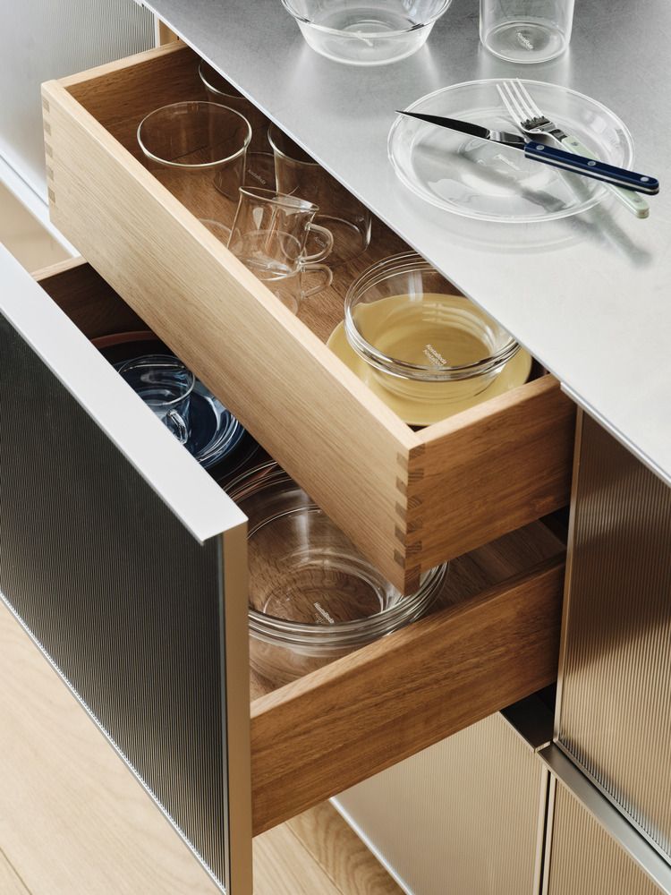 Öppna kökslådor i trä som visar olika glasföremål, med en metallbänk med en tallrik och bestick synlig ovanför.