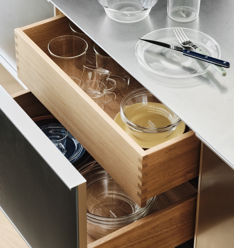 Öppna kökslådor i trä som visar olika glasföremål, med en metallbänk med en tallrik och bestick synlig ovanför.