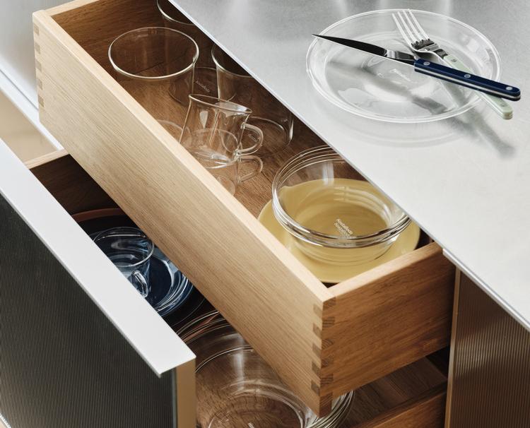 Öppna kökslådor i trä som visar olika glasföremål, med en metallbänk med en tallrik och bestick synlig ovanför.