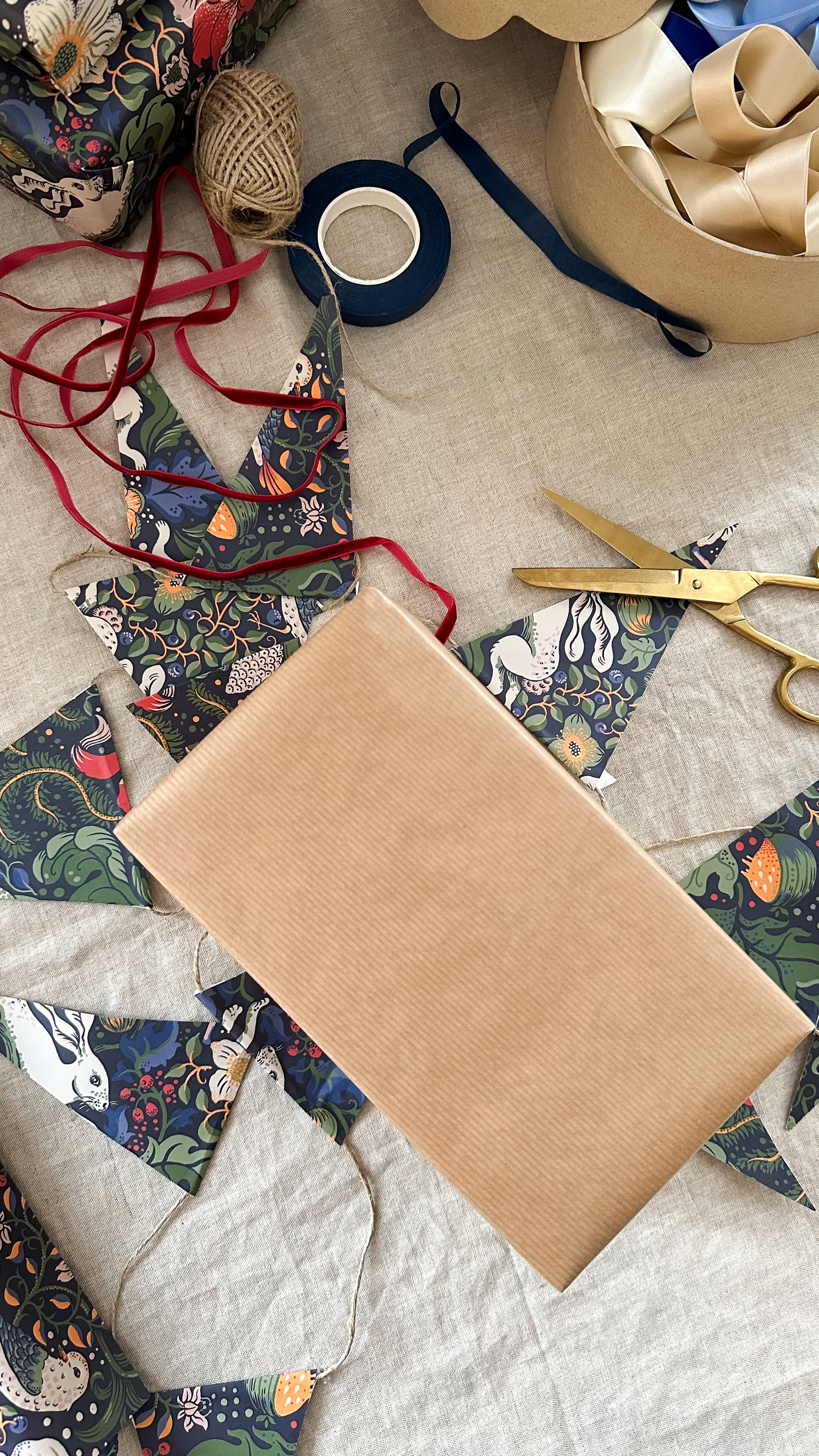 A gift wrapped in brown paper surrounded by patterned bunting, ribbons, twine, and gold scissors on a light fabric surface.