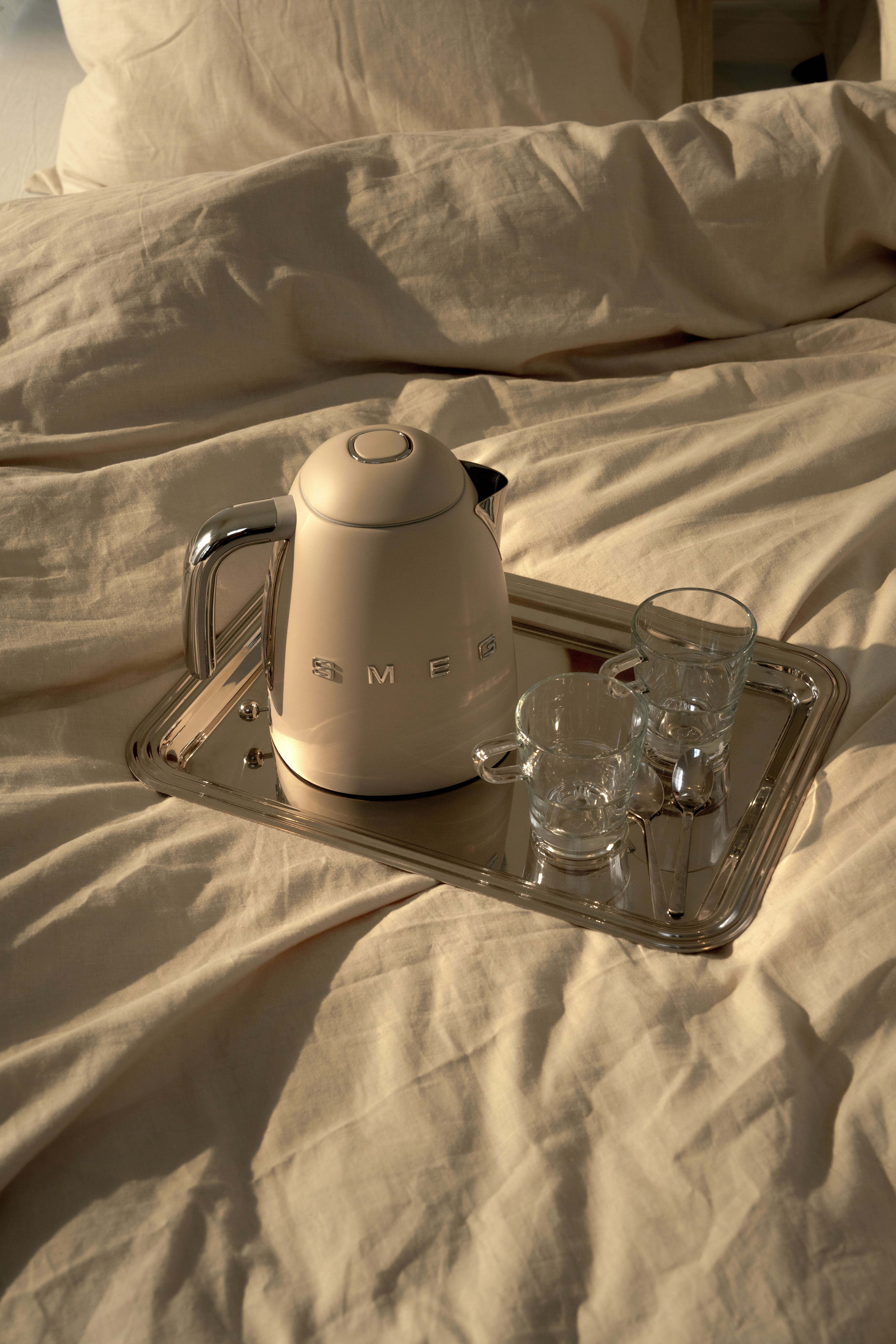 Beige Smeg-vattenkokare med två glas och skedar på en silverbricka i en solbelyst säng.