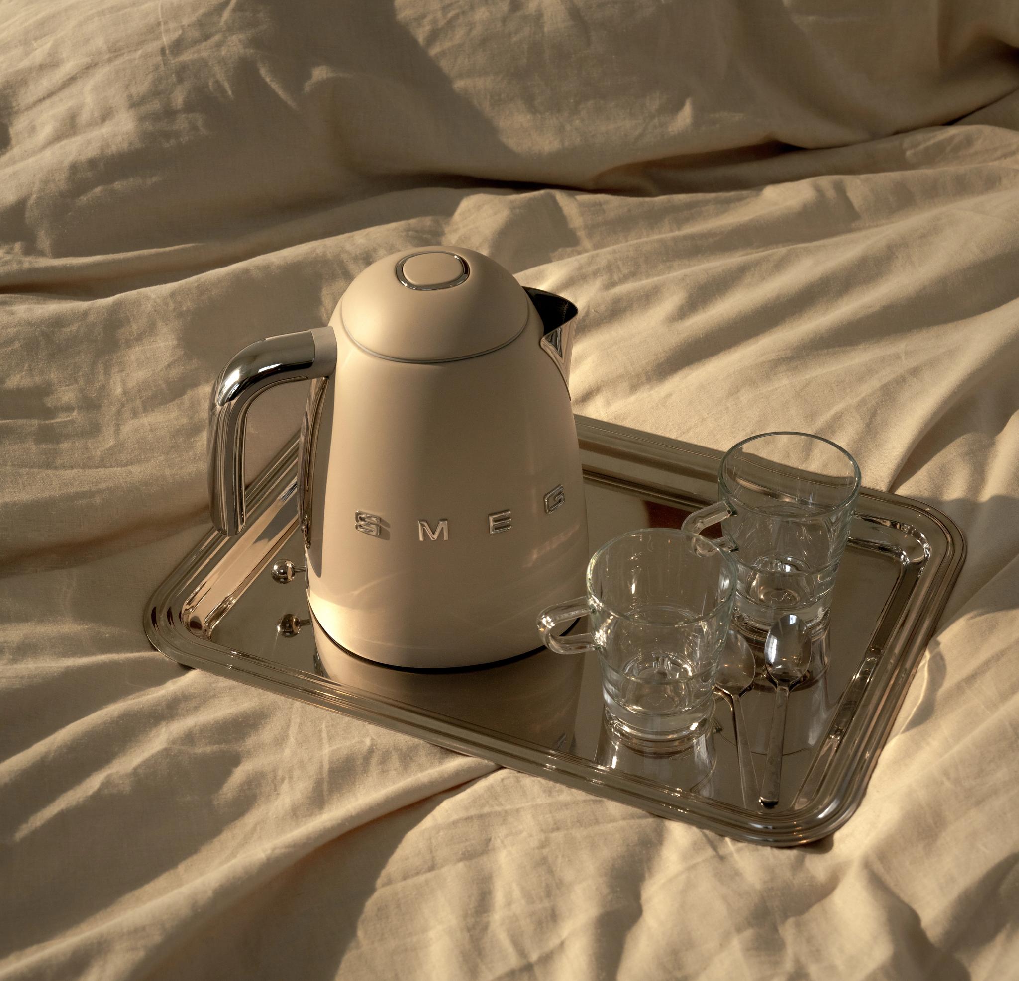 Beige Smeg-vattenkokare med två glas och skedar på en silverbricka i en solbelyst säng.