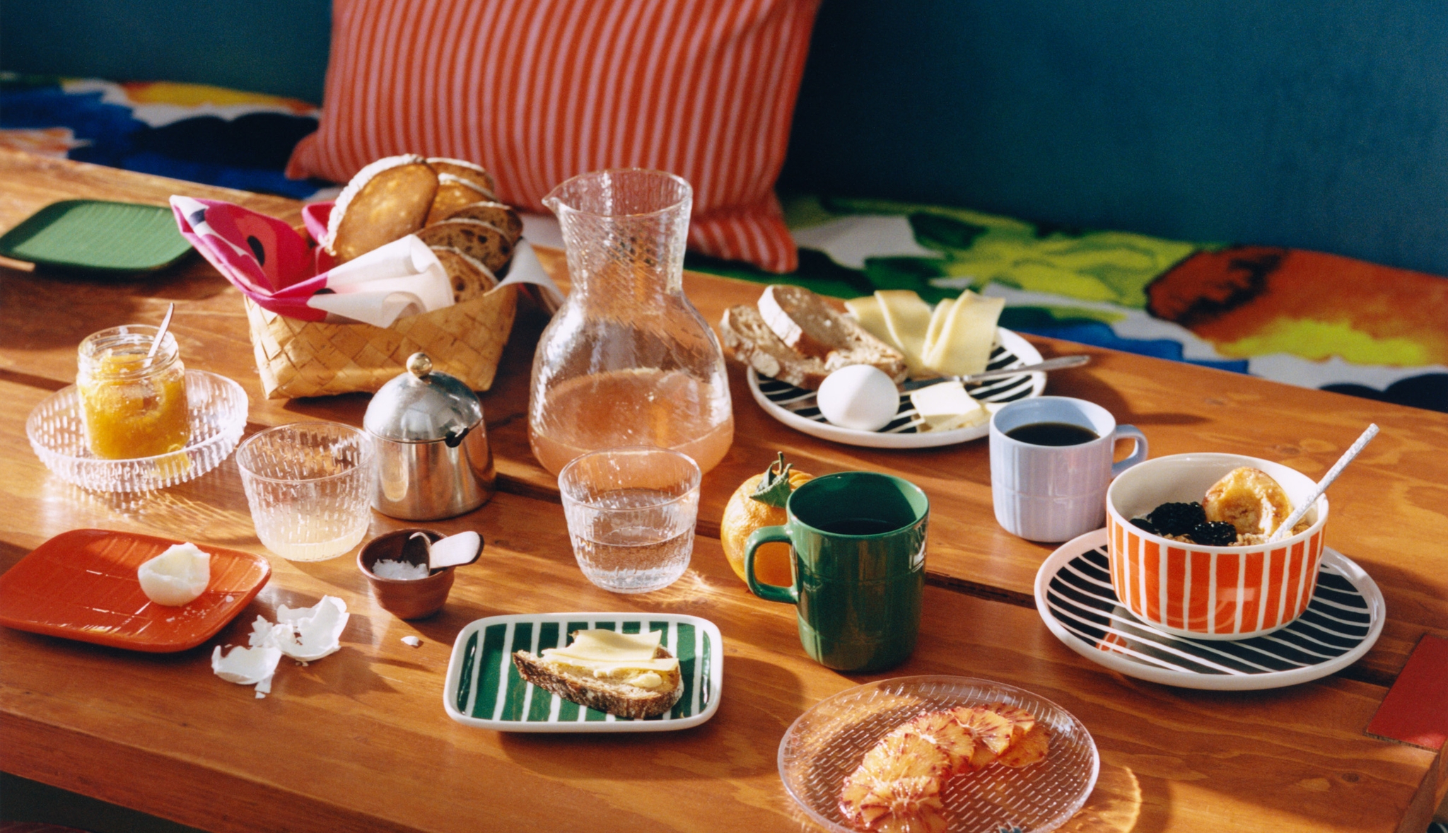 Fargerik frokostbordscene med brød, kaffe, juice og servise i sterke mønstrede farger på et trebord.