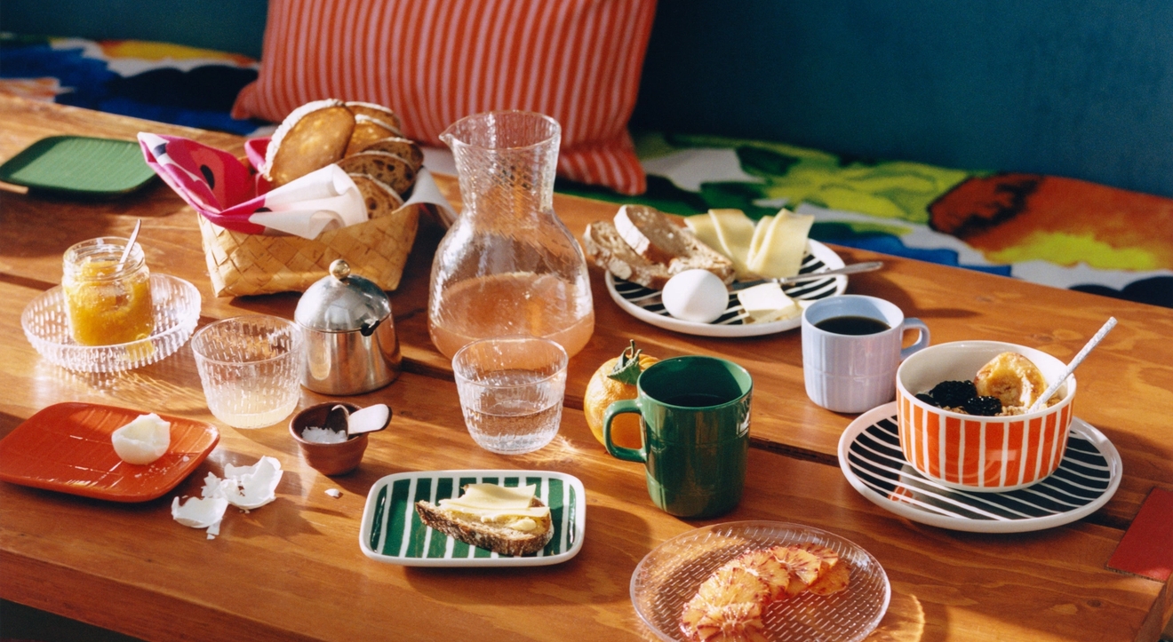 Fargerikt dekket frokostbord med brød, kaffe, juice og frukt på en trebenk med stripete puter og krakker.