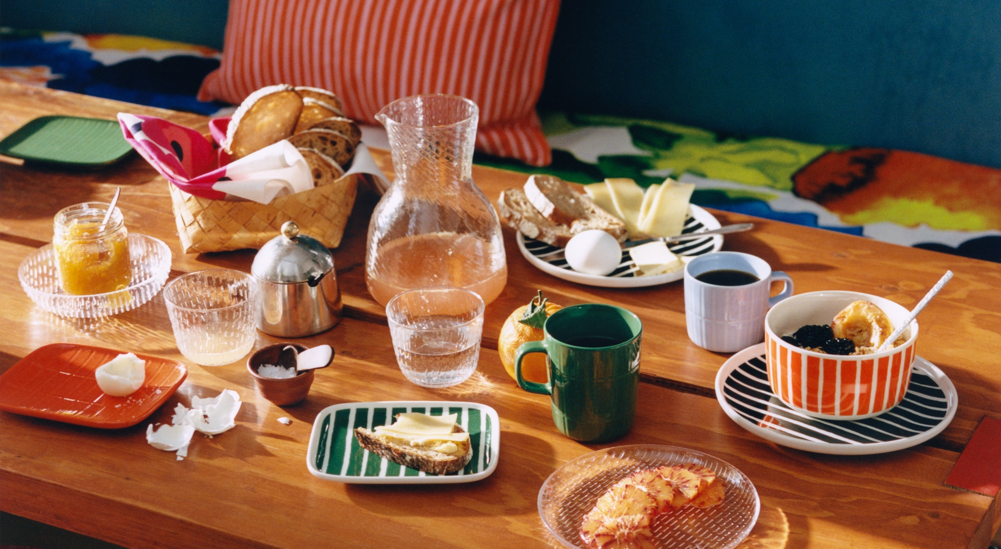Fargerikt dekket frokostbord med brød, kaffe, juice og frukt på en trebenk med stripete puter og krakker.