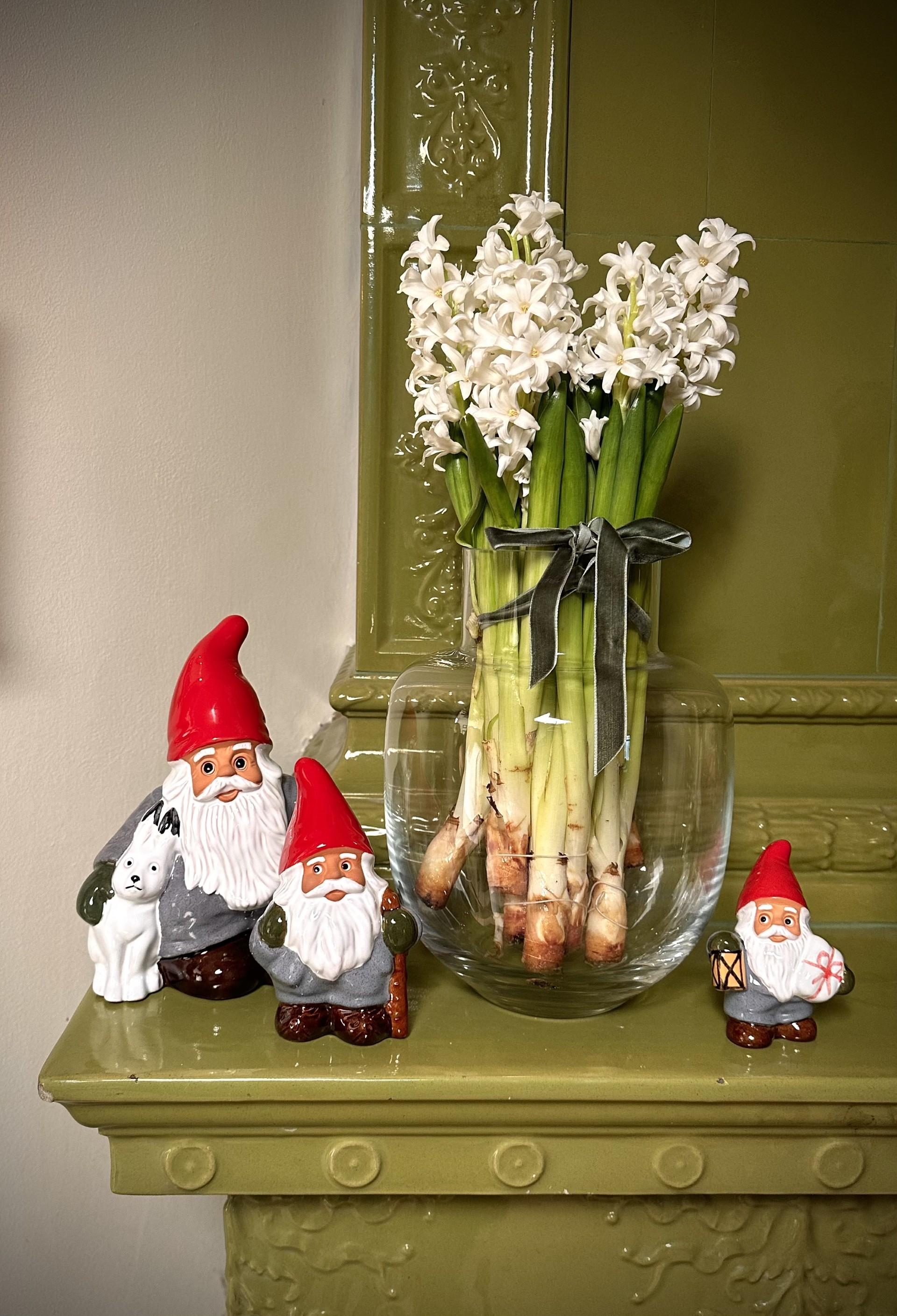 Kolme keraamista tonttua ja lasinen maljakko valkoisine hyasintteineen seisovat vihreällä, koristeellisella takanreunuksella.