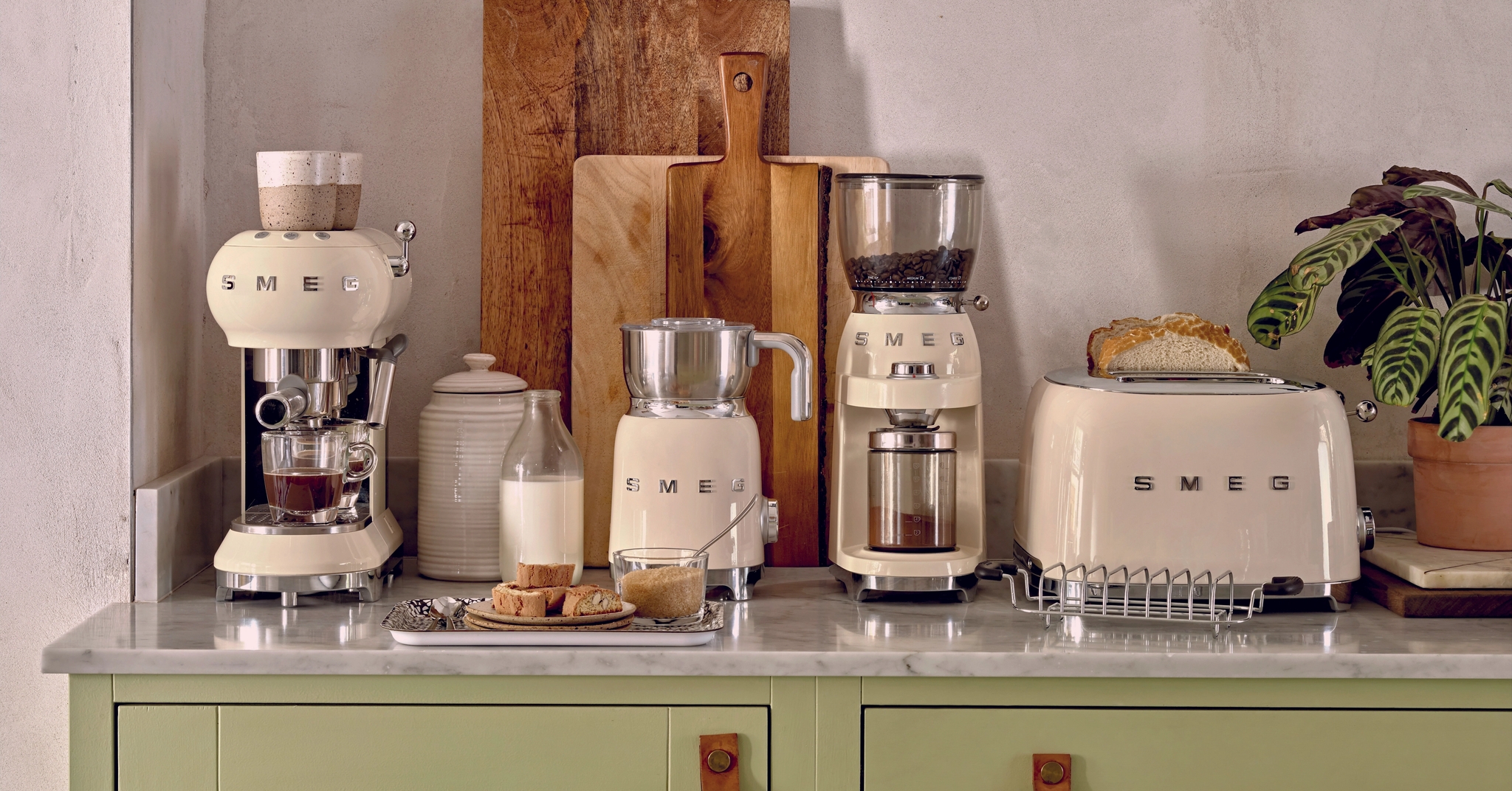 Stilren köksbänk med krämfärgade SMEG-köksmaskiner, brödrost, kaffemaskin och skärbrädor mot en ljus vägg.