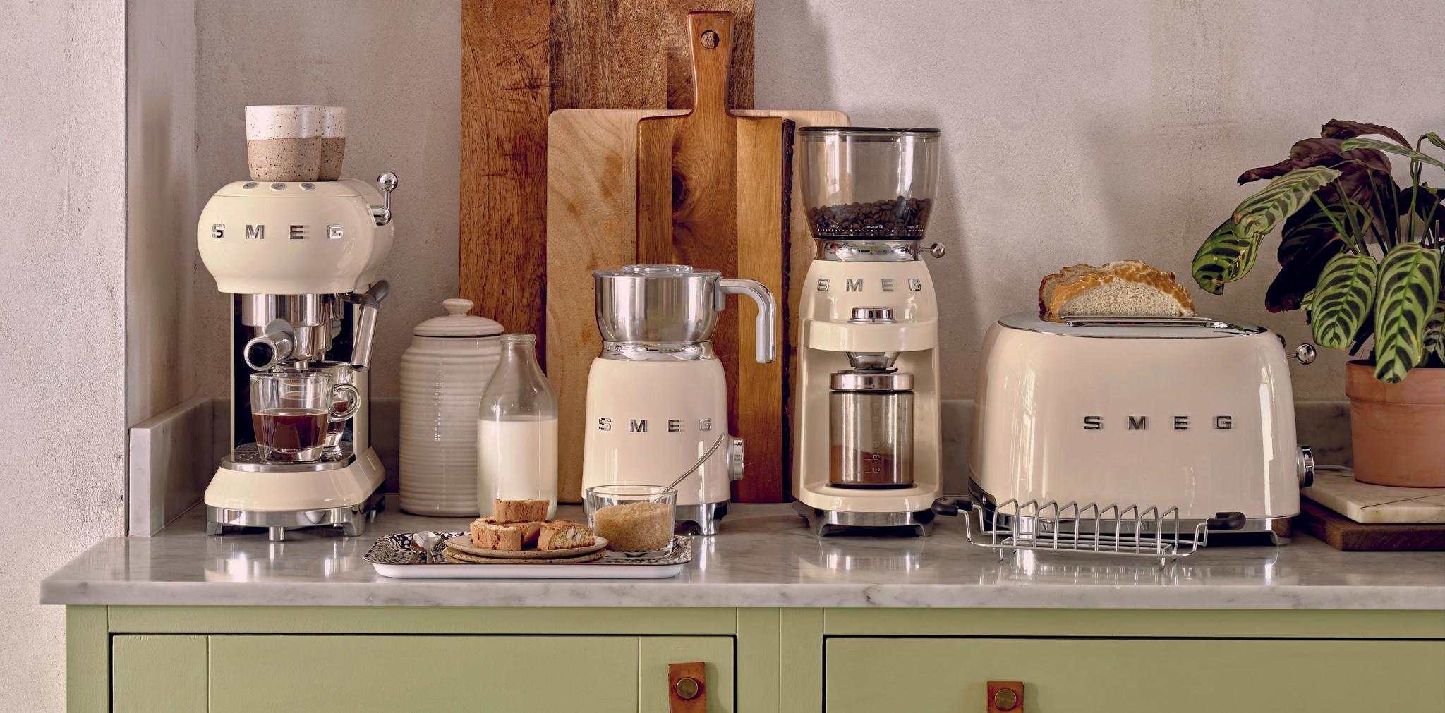Stilren köksbänk med krämfärgade SMEG-köksmaskiner, brödrost, kaffemaskin och skärbrädor mot en ljus vägg.