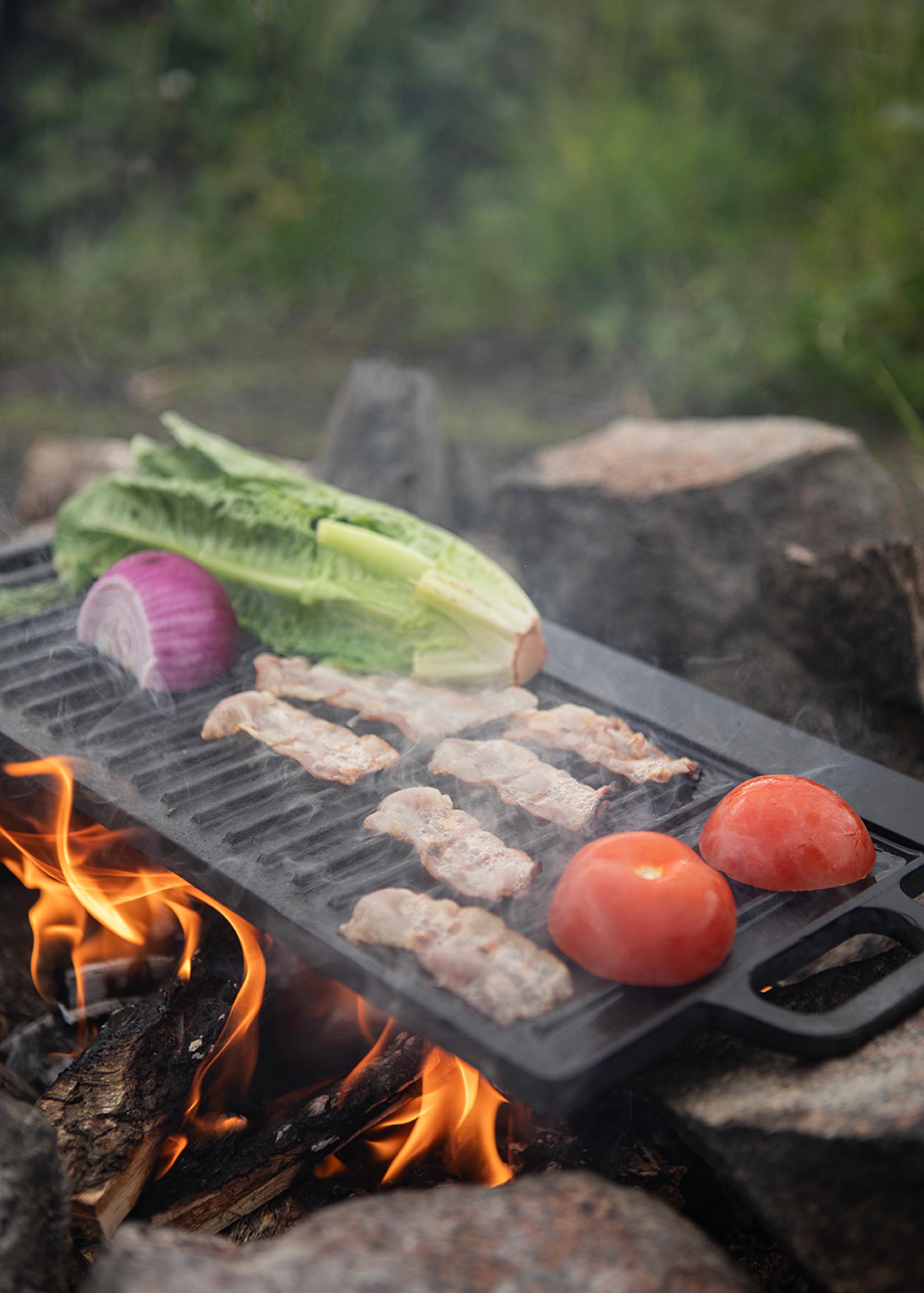 Grönsaker och bacon grillas över öppen eld på en gjutjärnsgrill utomhus.