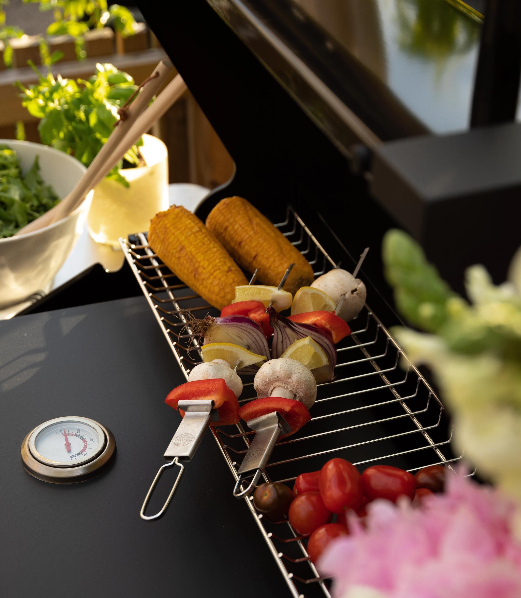 Mais, grønnsaksspyd og tomater på en grillrist ved siden av et termometer