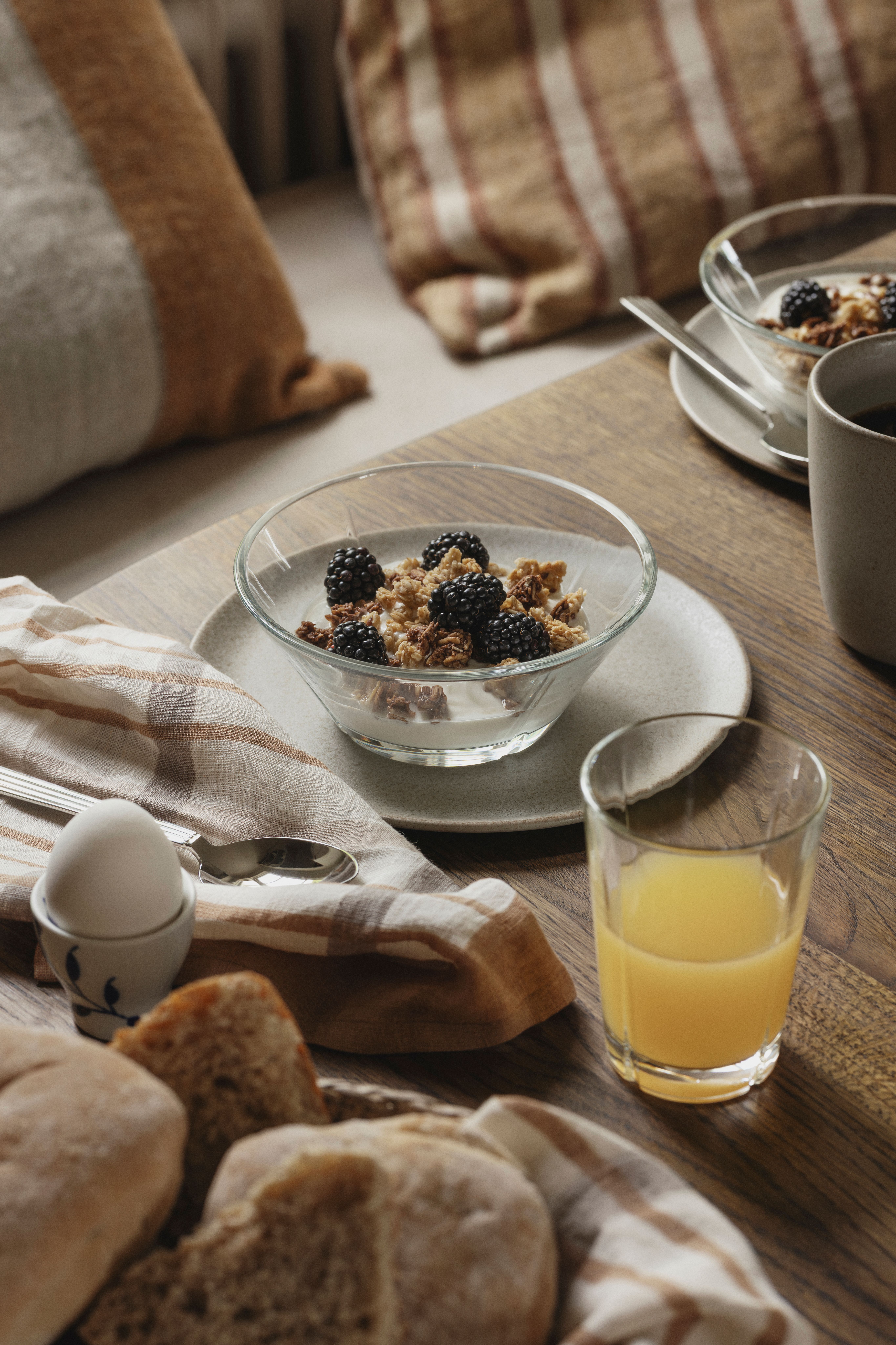 Rustik frukostdukning med yoghurt och granola med björnbär, nybakat bröd, kokt ägg och ett glas apelsinjuice på ett träbord.
