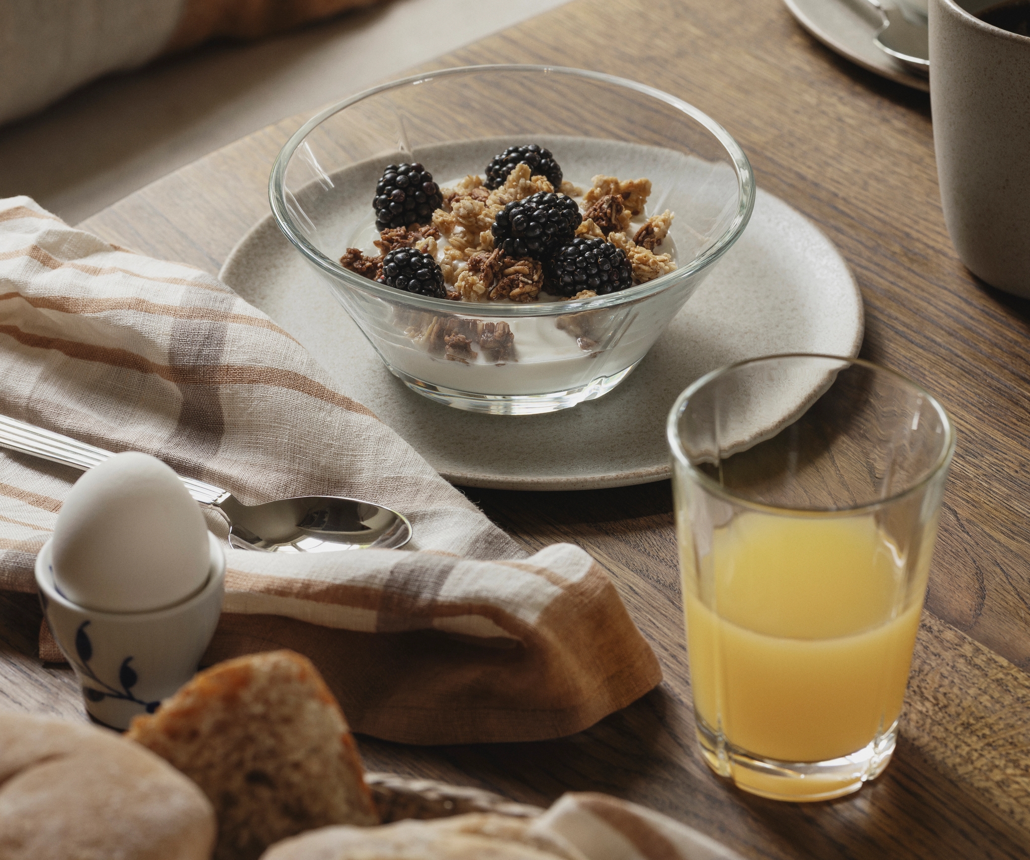 Rustik frukost med yoghurt och granola med björnbär, färskt bröd, kokt ägg och apelsinjuice på ett träbord.