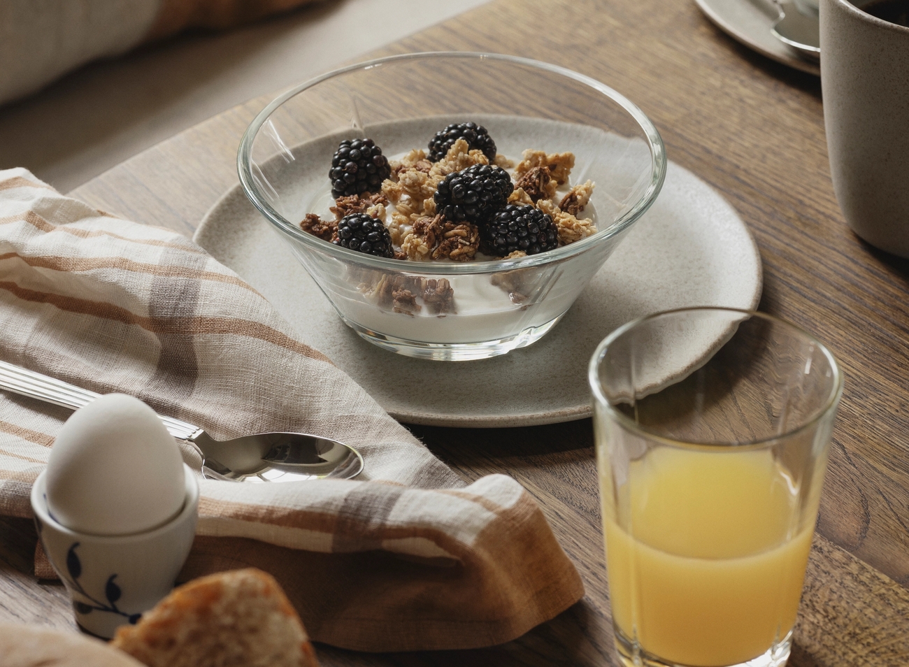 Rustik frukost med yoghurt och granola med björnbär, färskt bröd, kokt ägg och apelsinjuice på ett träbord.