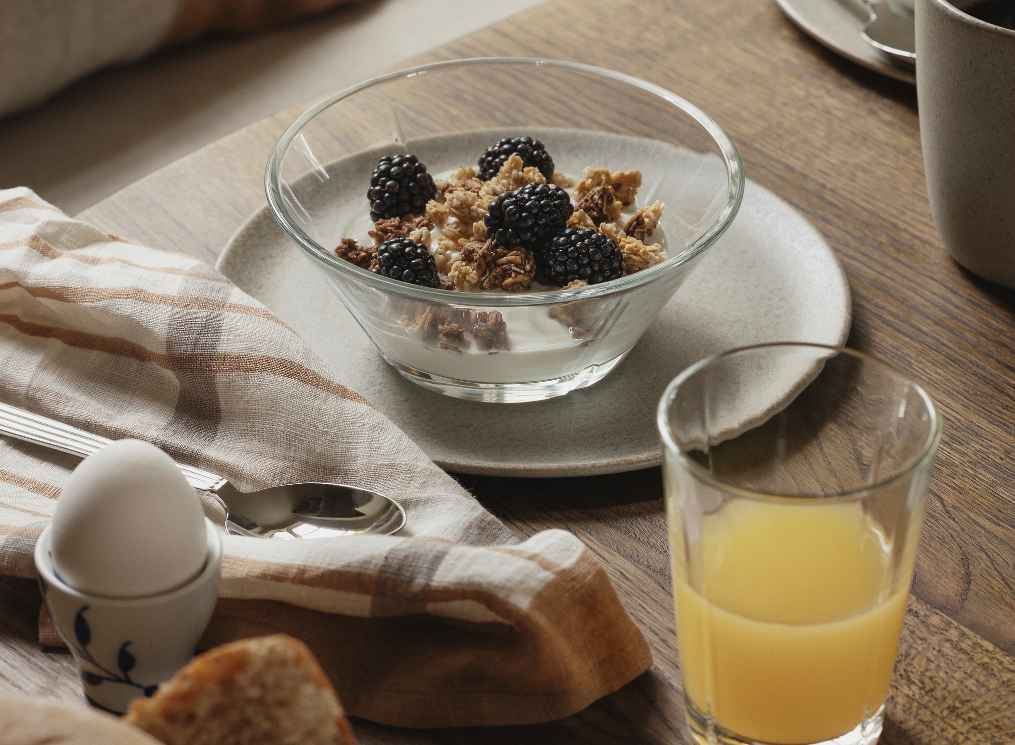 Rustik frukost med yoghurt och granola med björnbär, färskt bröd, kokt ägg och apelsinjuice på ett träbord.
