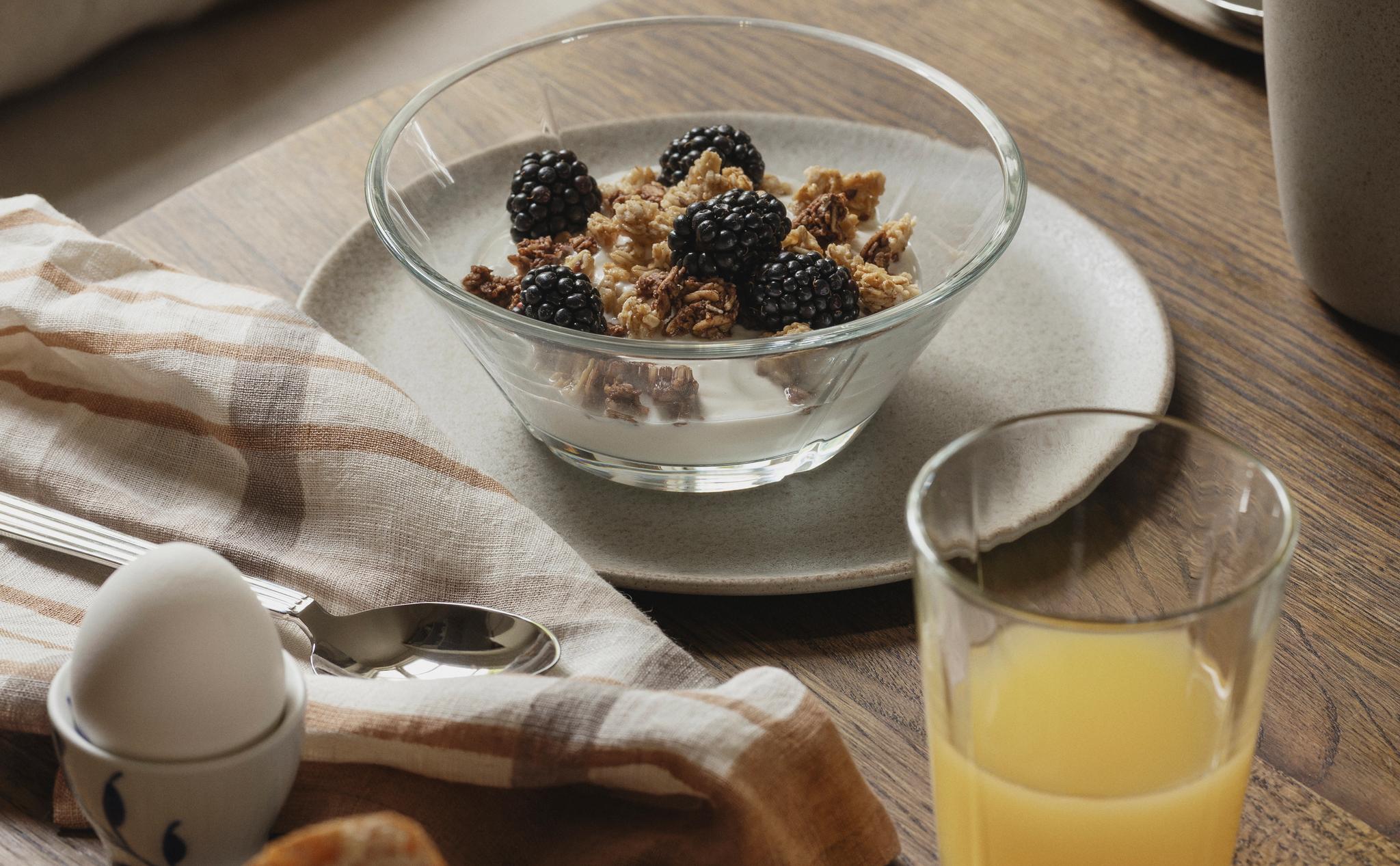 Rustik frukostdukning med yoghurt och granola med björnbär, nybakat bröd, kokt ägg och ett glas apelsinjuice på ett träbord.