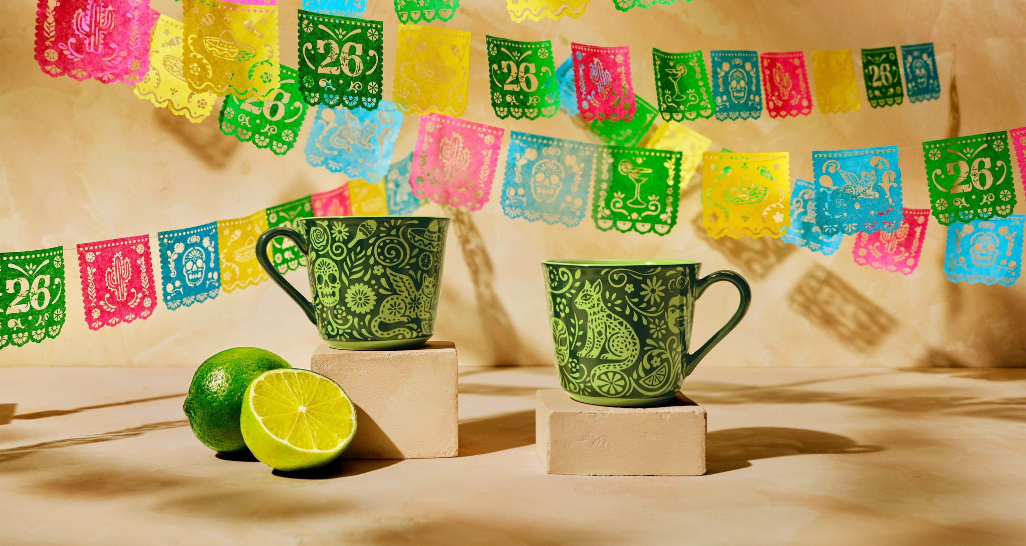 Två gröna, mexikanskt mönstrade koppar med limefrukter framför färgglada papel picado-girlanger.