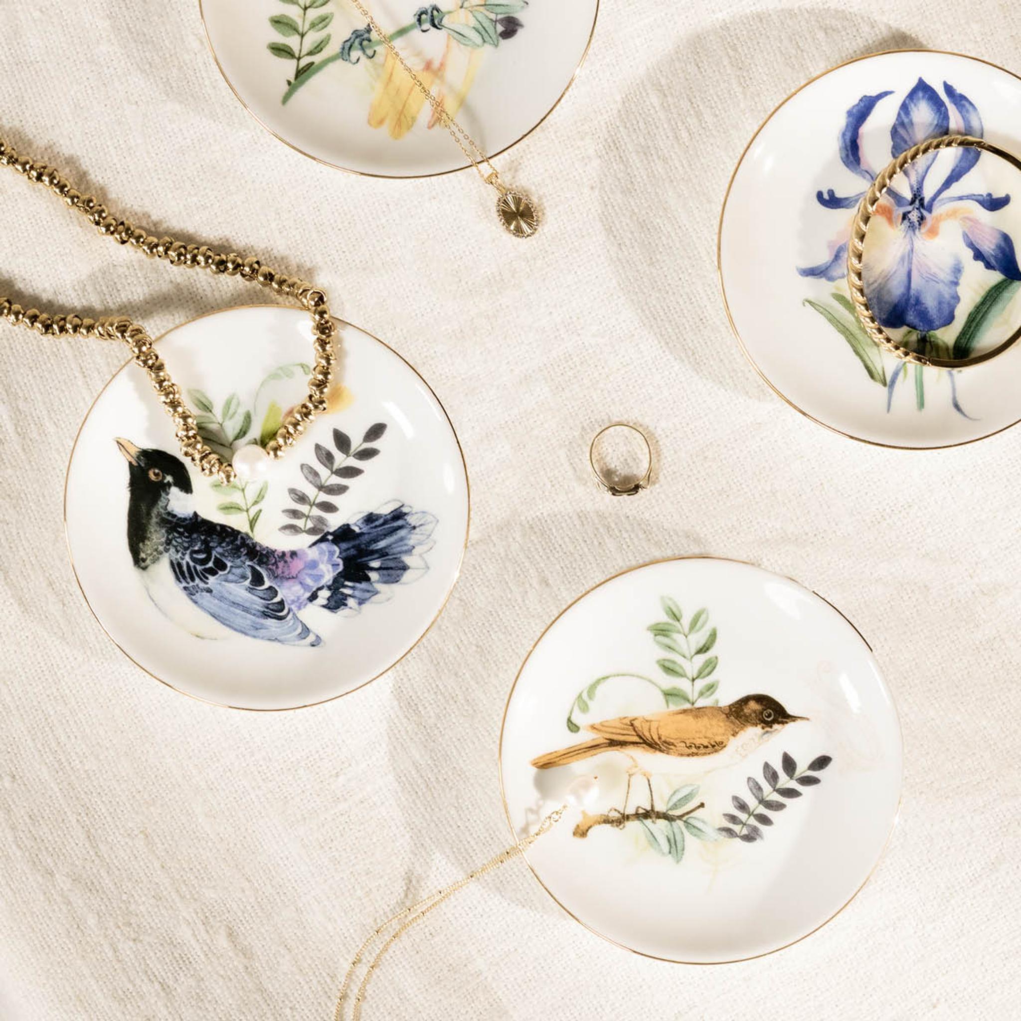 Fire hvite pynteskåler med gullkant og naturillustrasjoner holder halskjeder, en ring og et armbånd på et teksturert stoff.