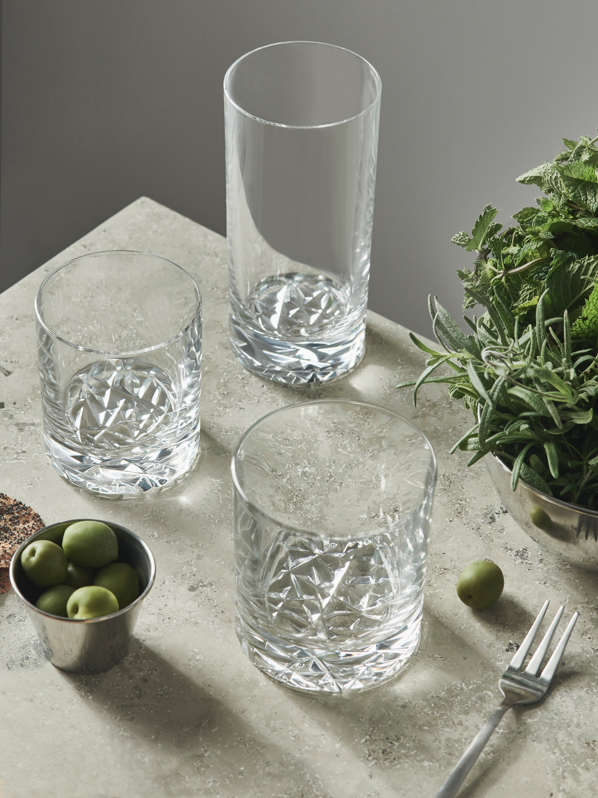 Tre mønstrede glass, en bolle med grønne oliven og friske urter på en grå overflate.