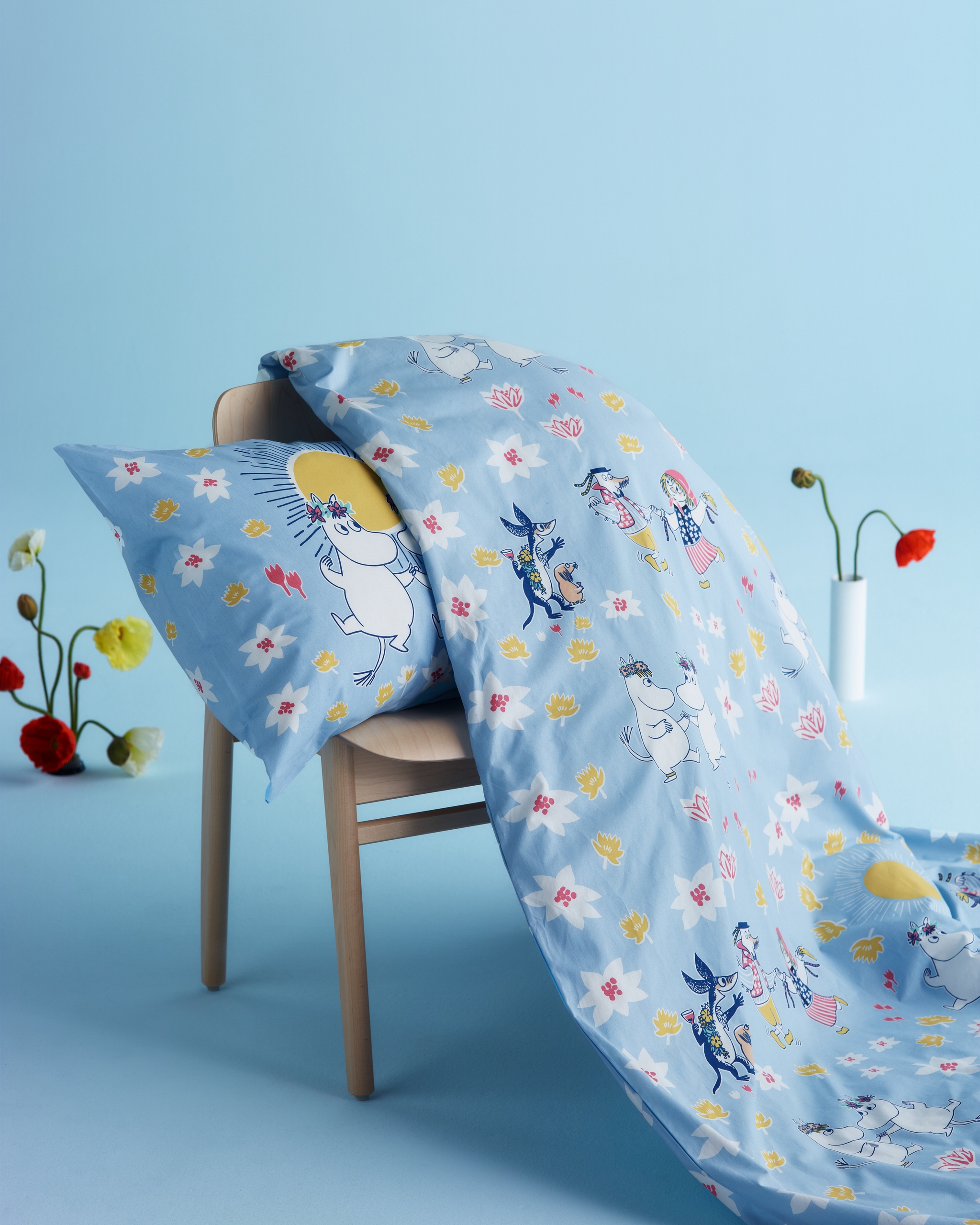 Blått påslakan och örngott med Mumin-motiv och blommor draperat över en trästol mot ljusblå bakgrund.