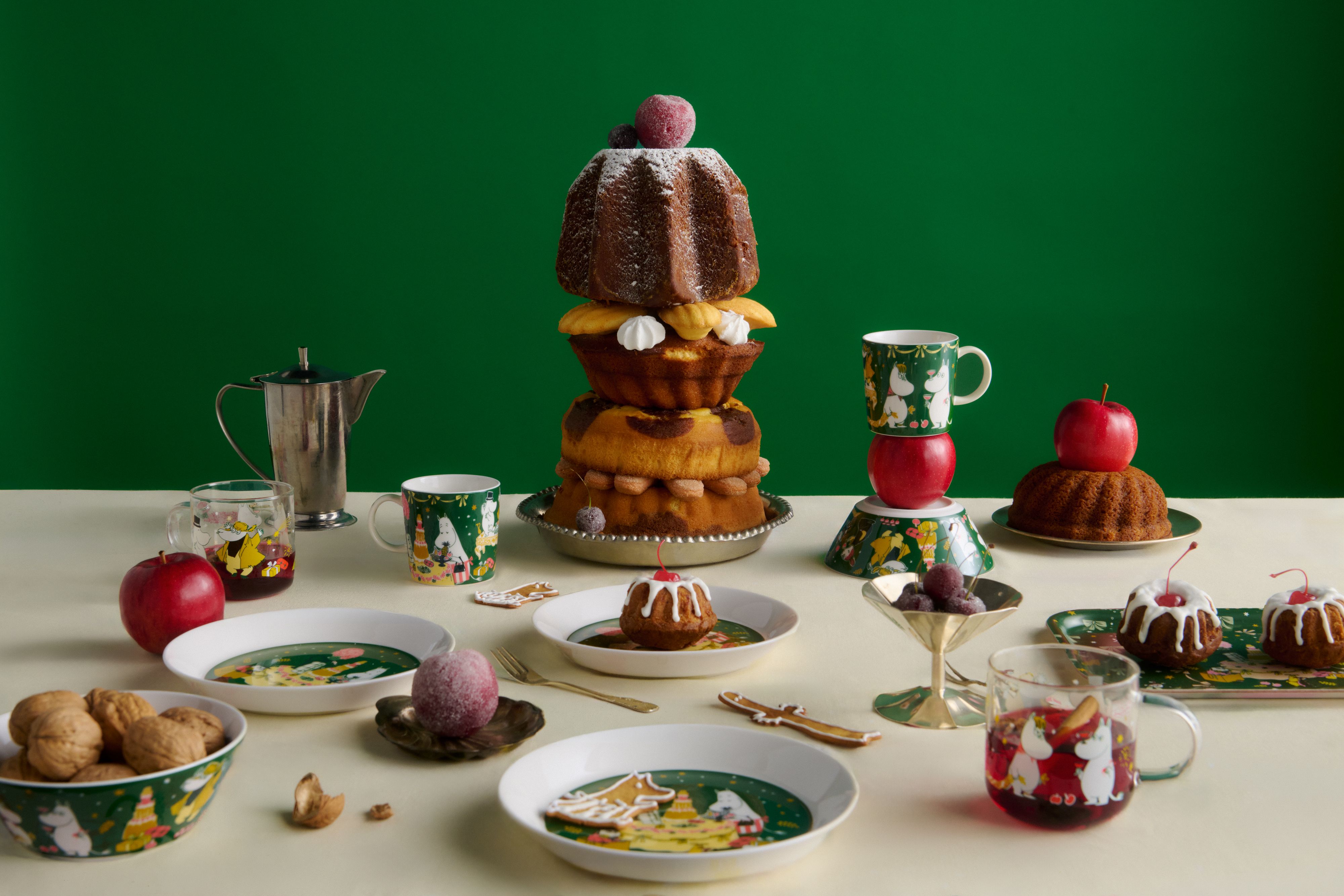 Festligt julbord med staplade kakor, röda äpplen och grönt Muminporslin mot en mörkgrön bakgrund.