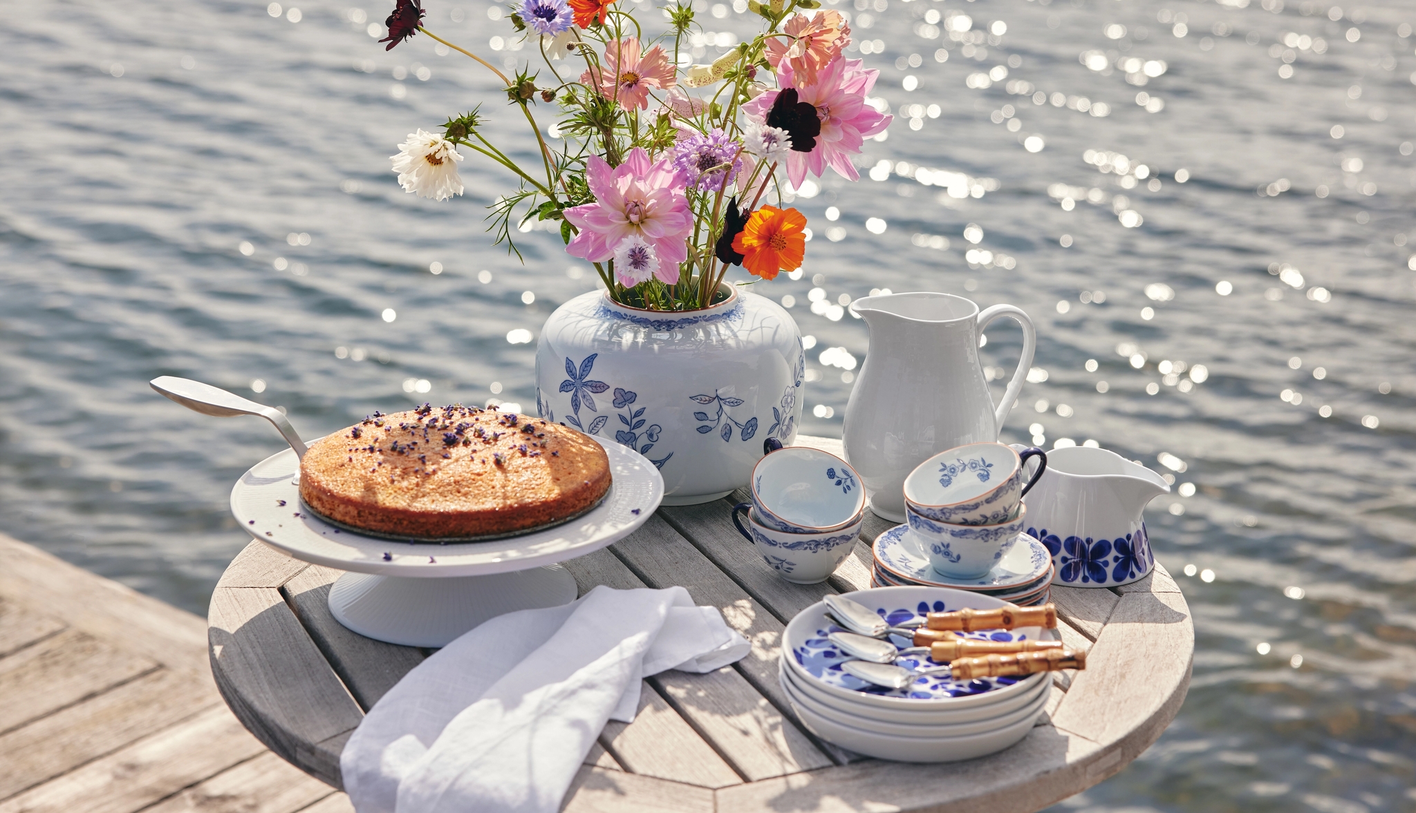 Somrig fikastund med tårta, blommor och blåvitt porslin på ett bord vid vattnet.