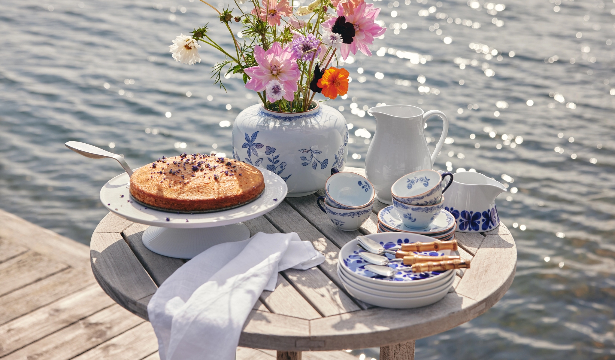 Somrig fikastund med tårta, blommor och blåvitt porslin på ett bord vid vattnet.