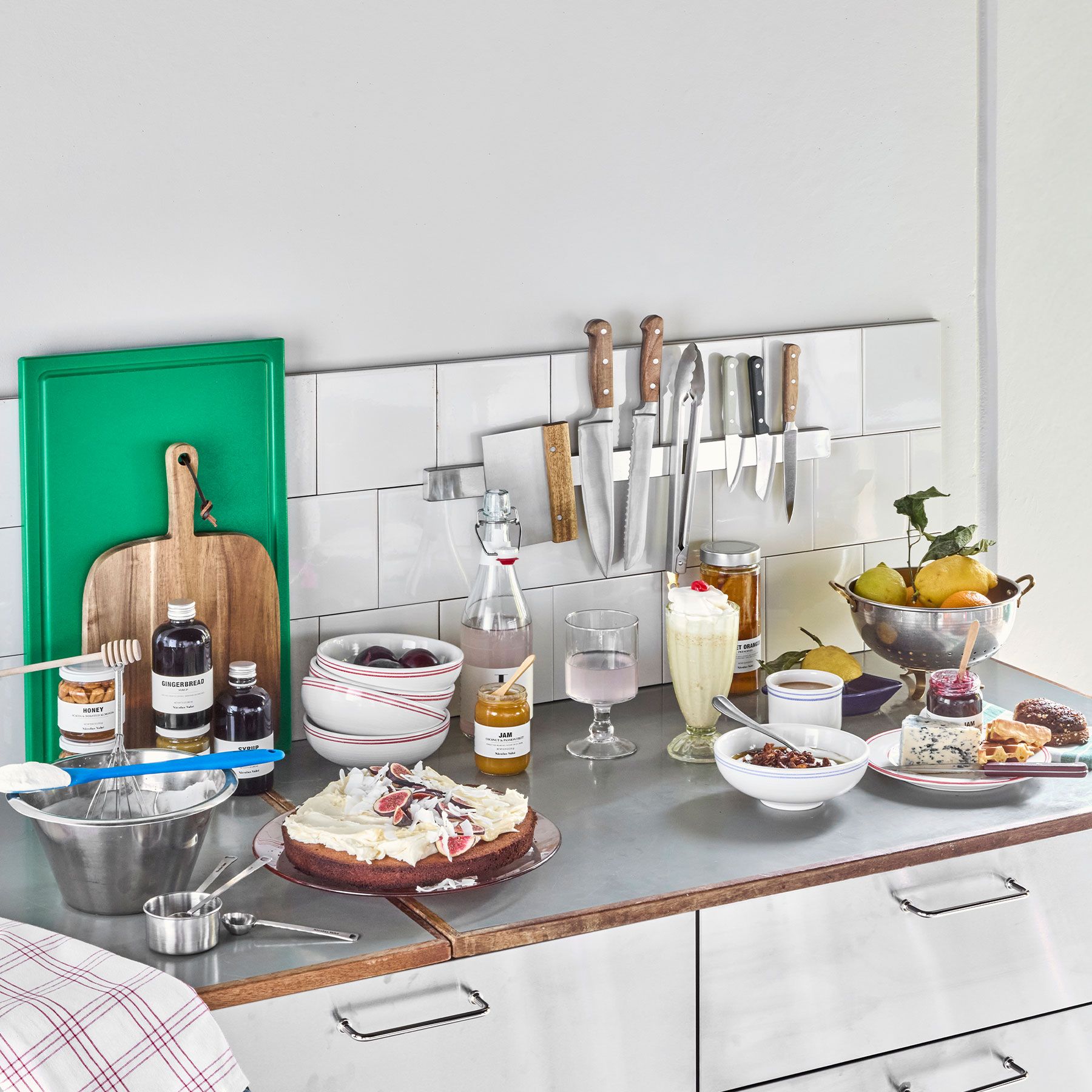 En köksbänk full av mat, inklusive en dekorerad tårta, färsk frukt, drycker och bakingredienser.