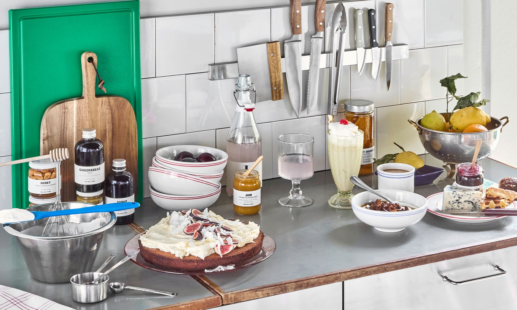 En köksbänk full av mat, inklusive en dekorerad tårta, färsk frukt, drycker och bakingredienser.
