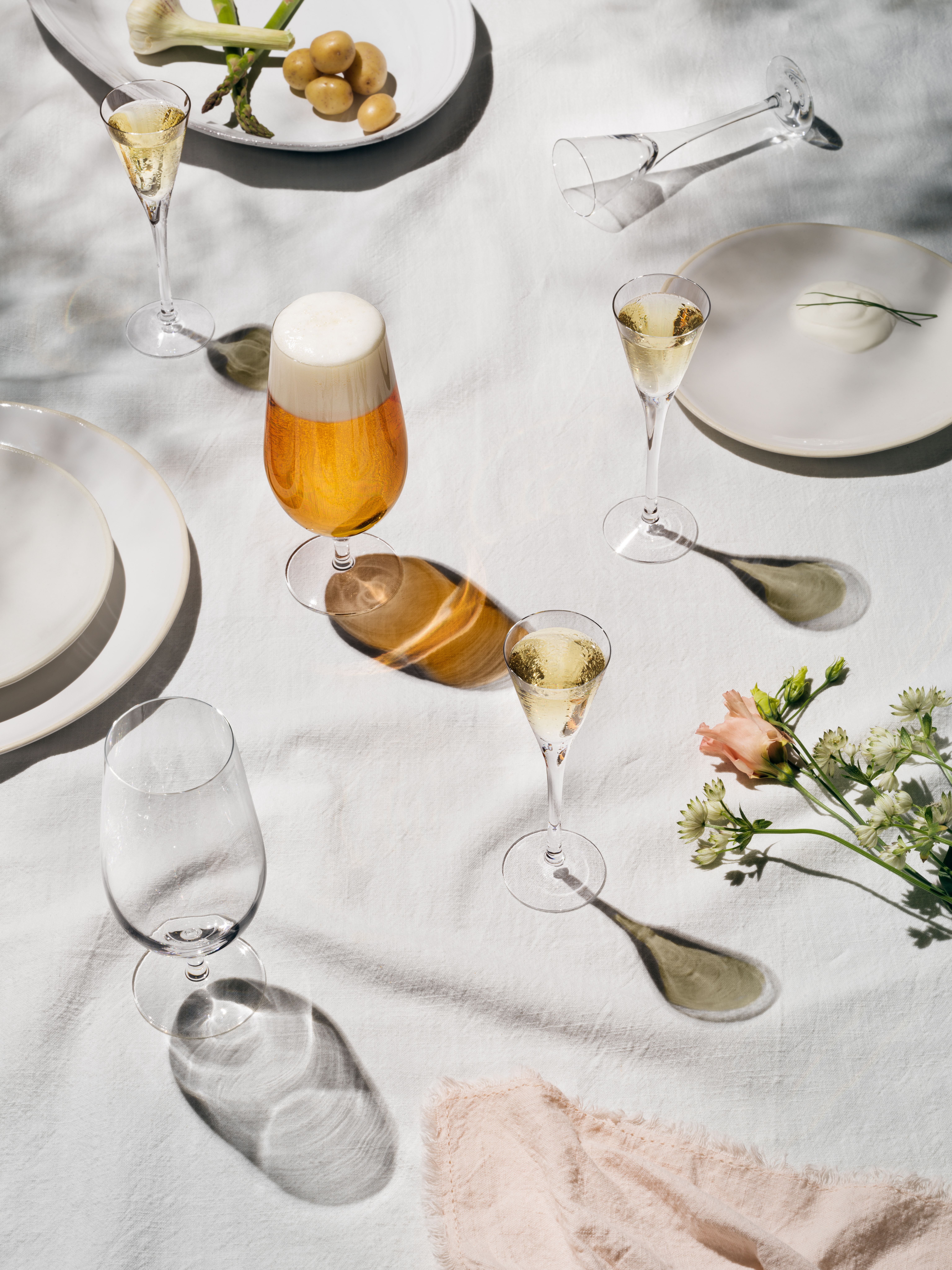 Somrig dukning med öl, mousserande vin, potatis och blommor på vit linneduk i mjukt solljus.