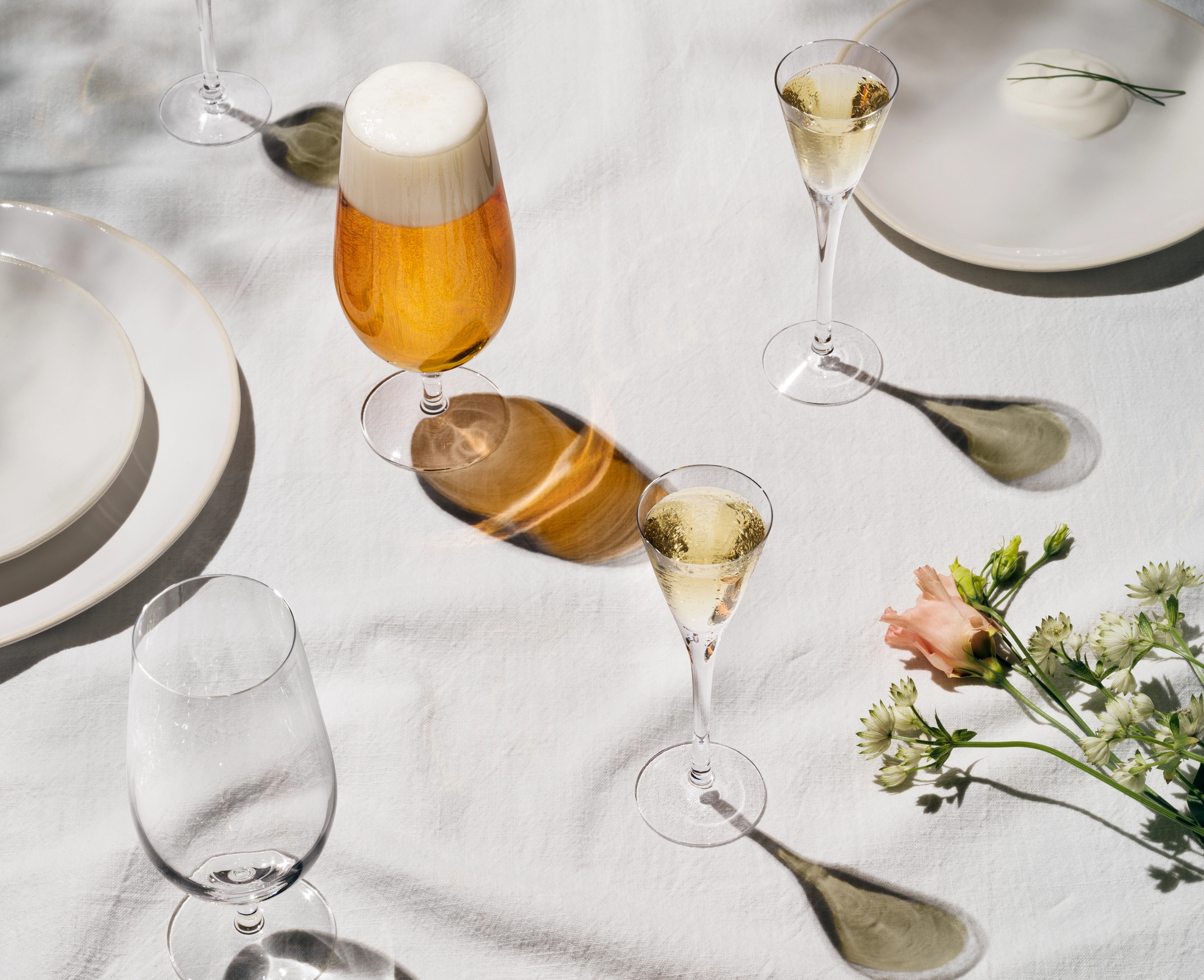 Elegant dukning med öl, mousserande vin, potatis och vårprimörer på vit linneduk i mjukt solljus.