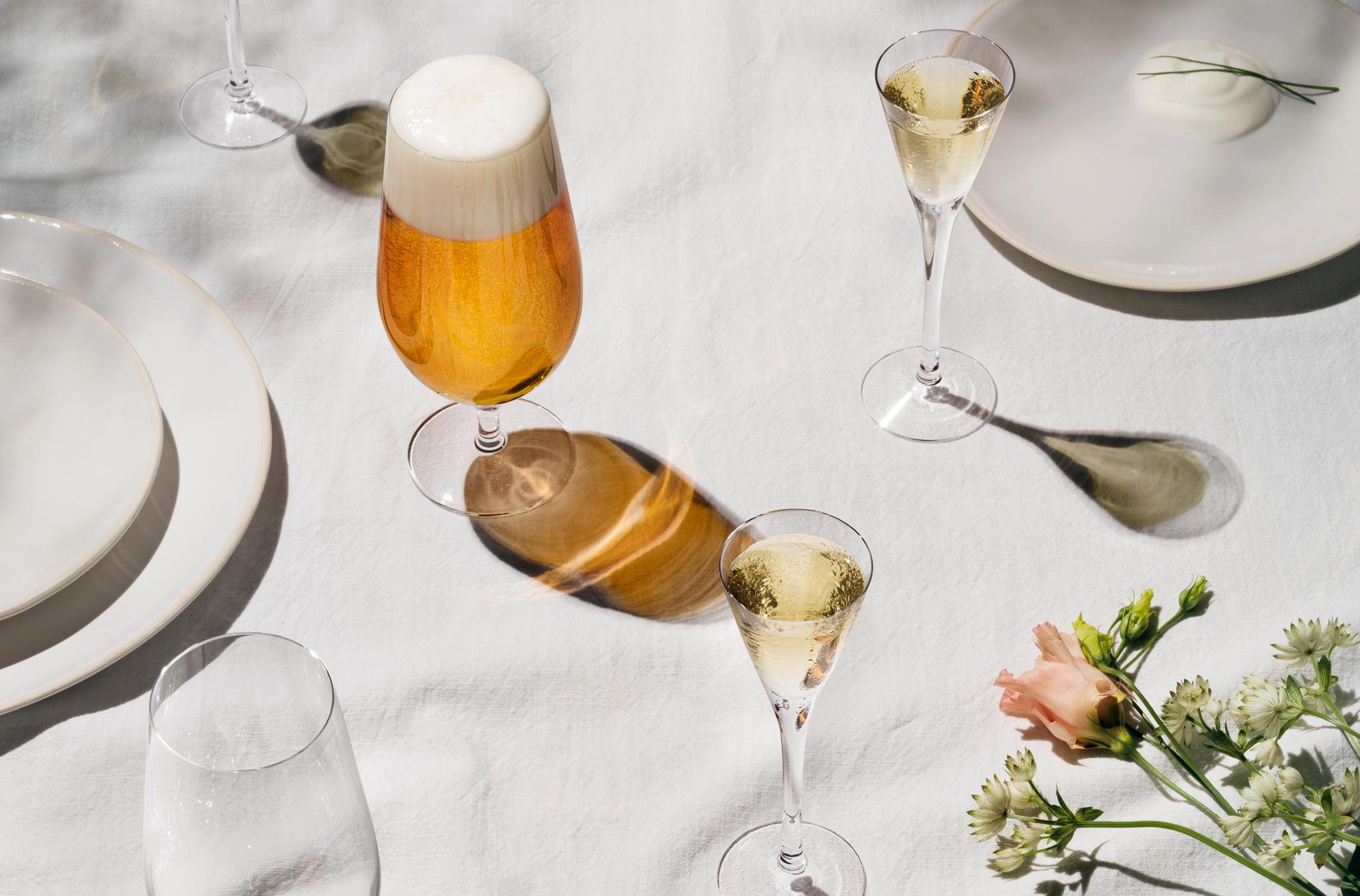 Somrig dukning med öl, mousserande vin, potatis och blommor på vit linneduk i solljus.